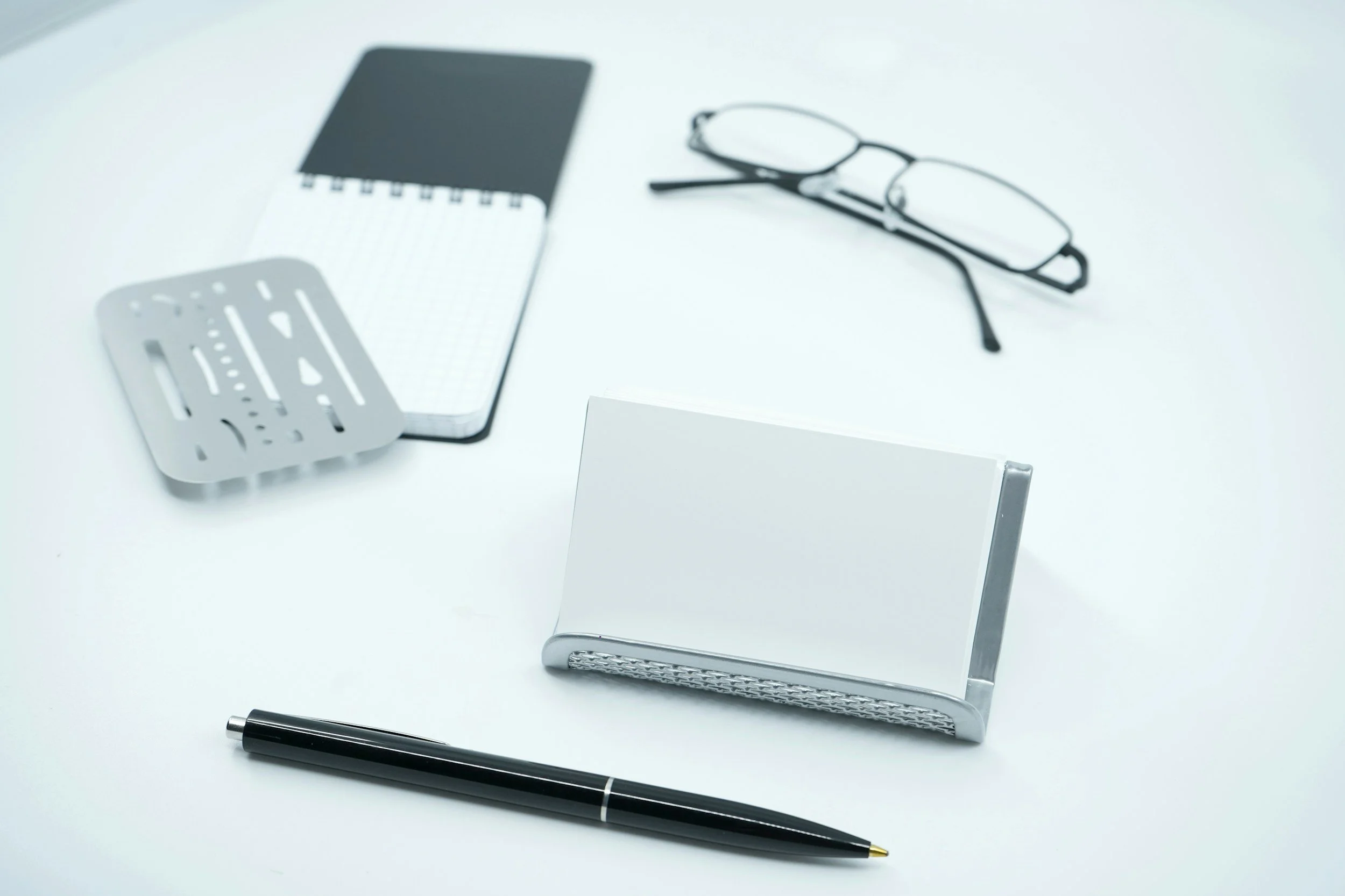 A desk containing a black pen, a mirror with a textured silver frame, a small silver notepad holder, a pair of black eyeglasses, a spiral notepad with black cover, and a white stand with a blank card.