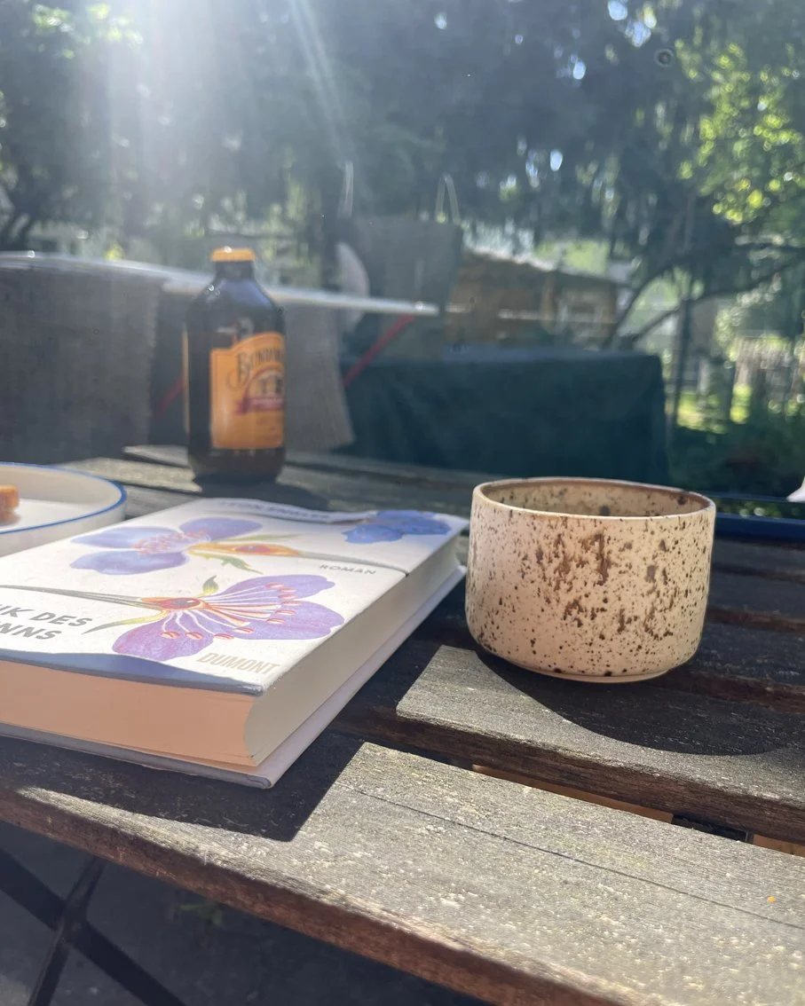 Ein Holztisch im Freien mit einem Buch, einer gesprenkten handgemachten Keramiktasse, einer Flasche und einem Teller im Sonnenlicht, umgeben von Grün.