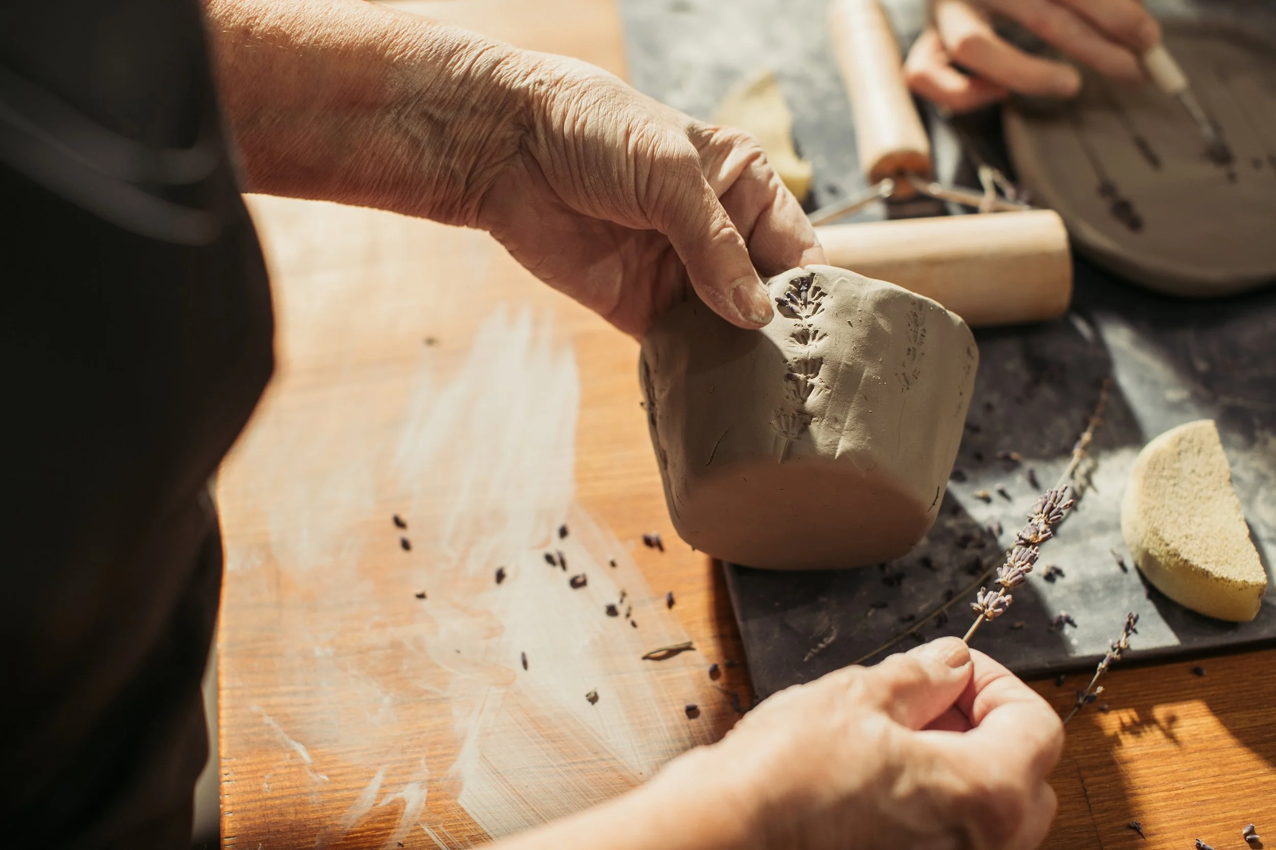 Hand einer Person, die eine Tonfigur bearbeitet, auf einem Holzbrett mit Werkzeugen und weiteren Tonfiguren.