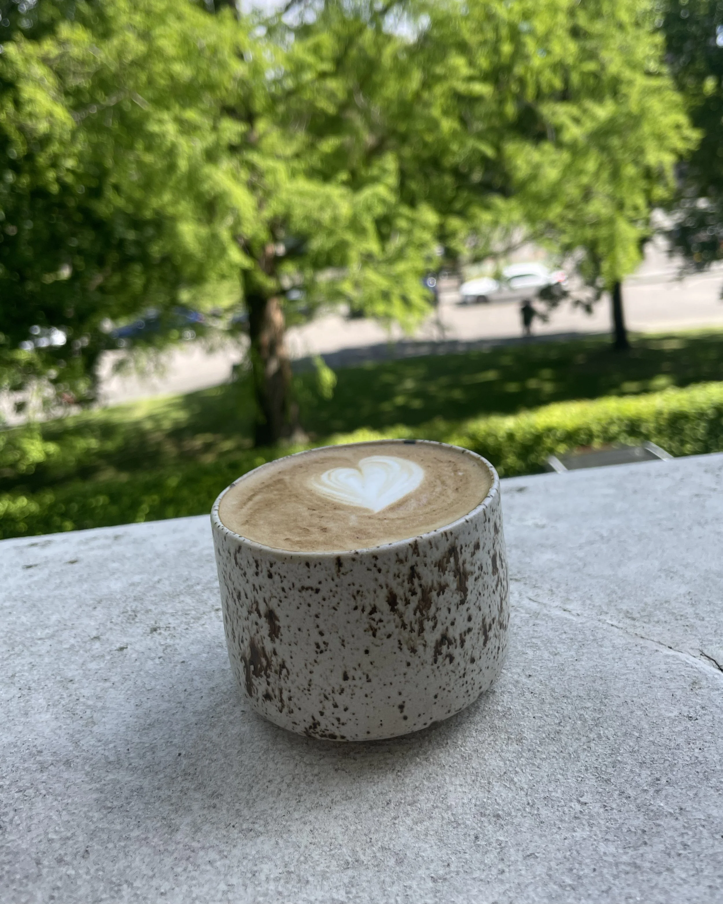 Hangemachter Keramik-Becher mit Latte-Art auf einem weißen Tisch im Freien, im Hintergrund grüne Bäume und eine Straße mit Autos.