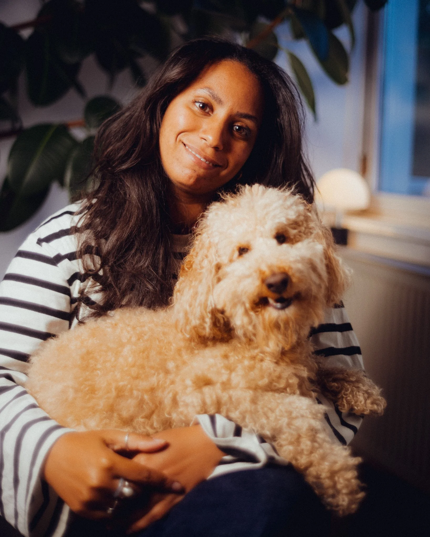 Eine Frau mit dunklen, langen Haaren hält einen goldgelben Hund, vermutlich ein Labradoodle, auf ihrem Schoß. Das Foto wurde in Innenräumen aufgenommen, mit Pflanzen im Hintergrund und einem Fenster auf der rechten Seite.