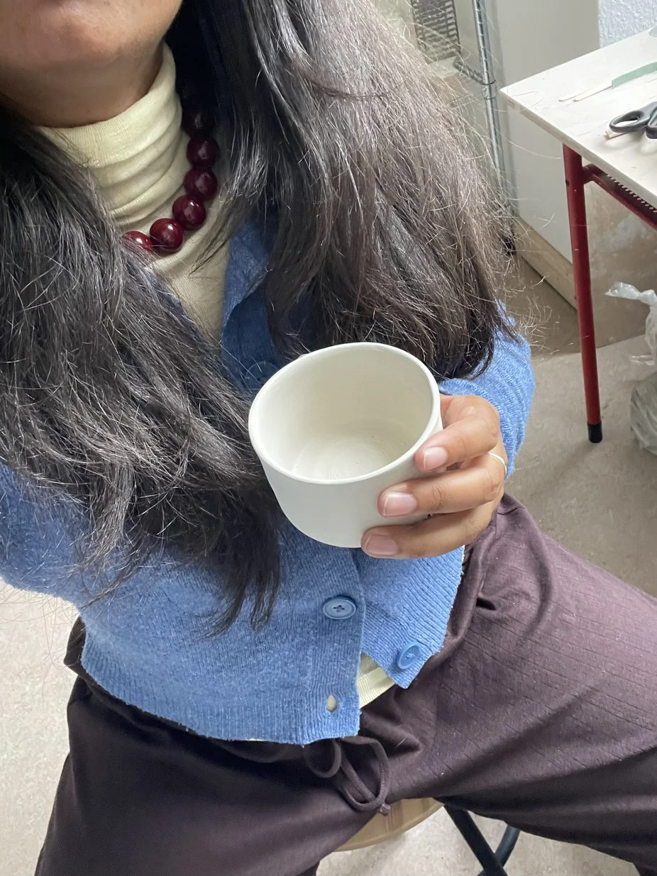 Eine Frau sitzt auf einem Stuhl, hält eine weiße handgemachte Keramik Tasse in der Hand, trägt eine cremefarbene Turtleneck, eine blaue Strickjacke, eine rote Perlenkette und braune Hosen in einem Raum mit Werkstatt ausgestattet.