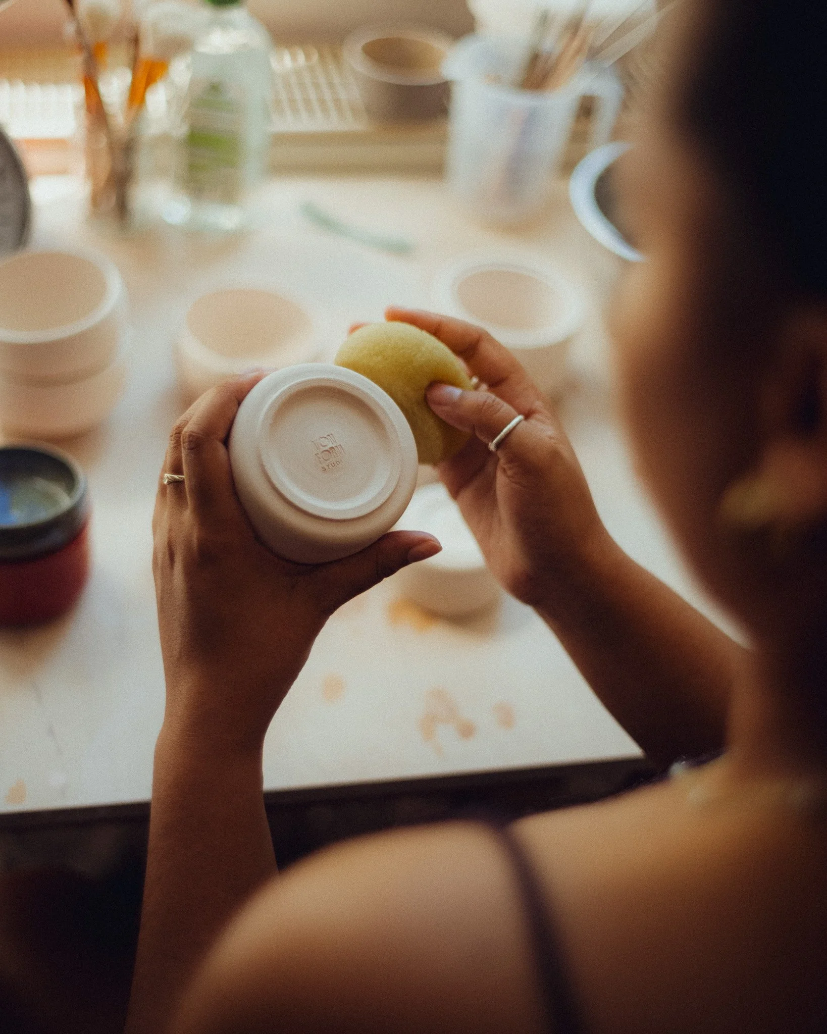 Nahaufnahme von Händen, die eine Tasse halten, auf einem Tisch mit handgemachten Bechern aus Keramik und verschiedenen Töpfer-Untensilien im Hintergrund.