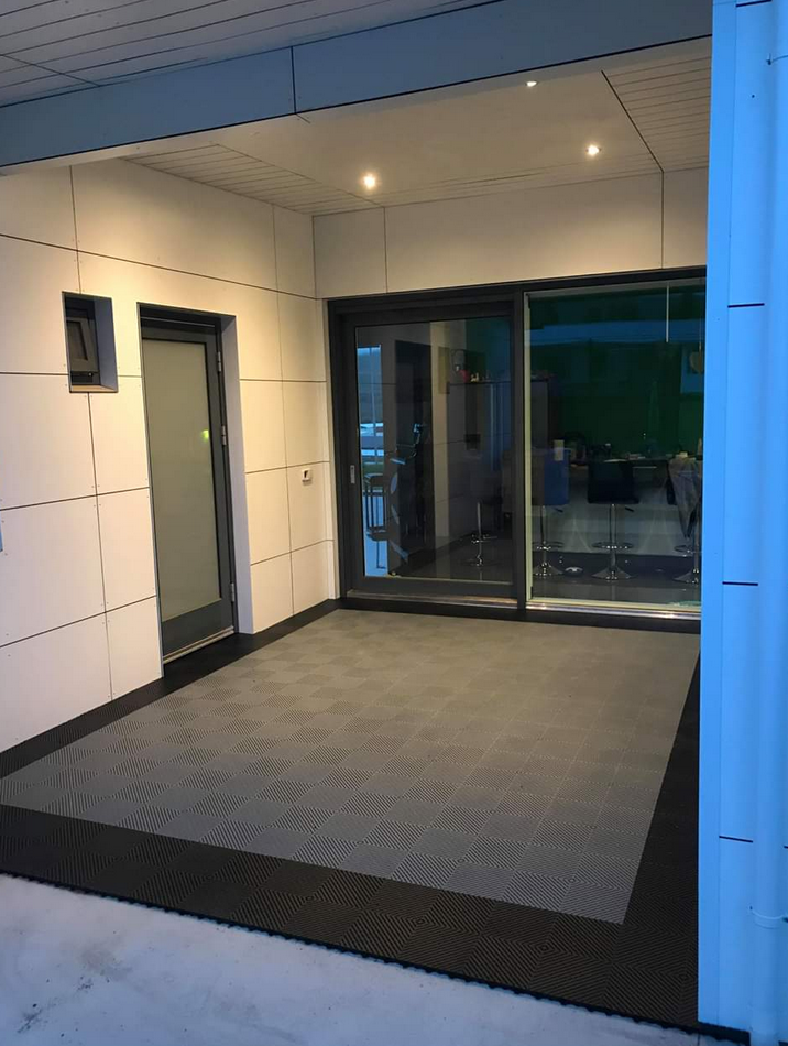 Empty balcony or patio area with glass door and windows, beige tiled walls, and textured Swisstrax tiles on the floor, inside a modern building.