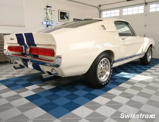 A vintage white muscle car with blue racing stripes, parked inside a garage with a checkered swisstrax tile floor.