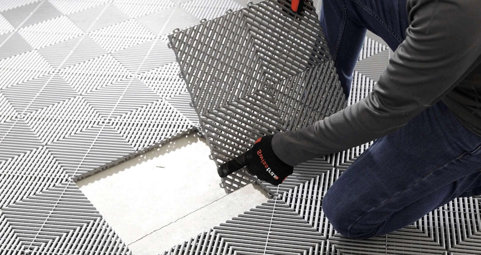 Person kneeling on ground while installing a gray swisstrax plastic tile flooring with a square grid pattern, wearing gloves and a black jacket.