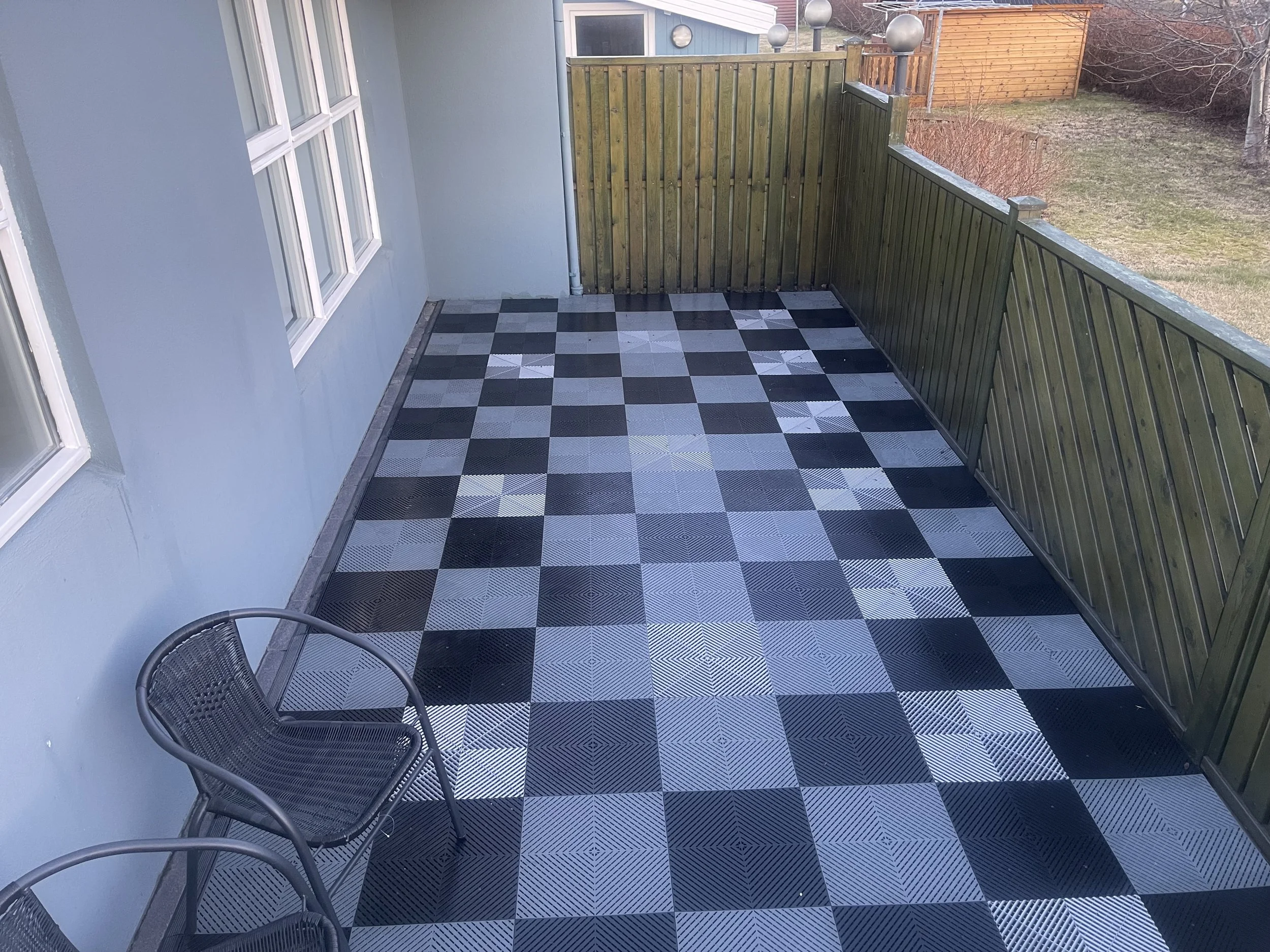 Empty balcony with black and gray checkered swisstrax flooring, two metal chairs in the corner, blue exterior wall with three white-framed windows, wooden fence, and outdoor yard with grass and trees in the background.