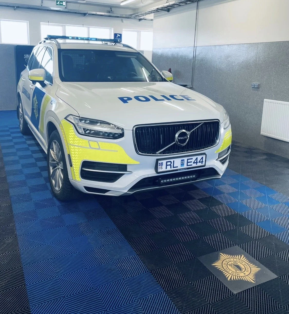A white police car with yellow and blue markings parked indoors, with a blue light bar on top. It is parked on a black and blue checkered swisstrax floor with a police star embedded in the floor.