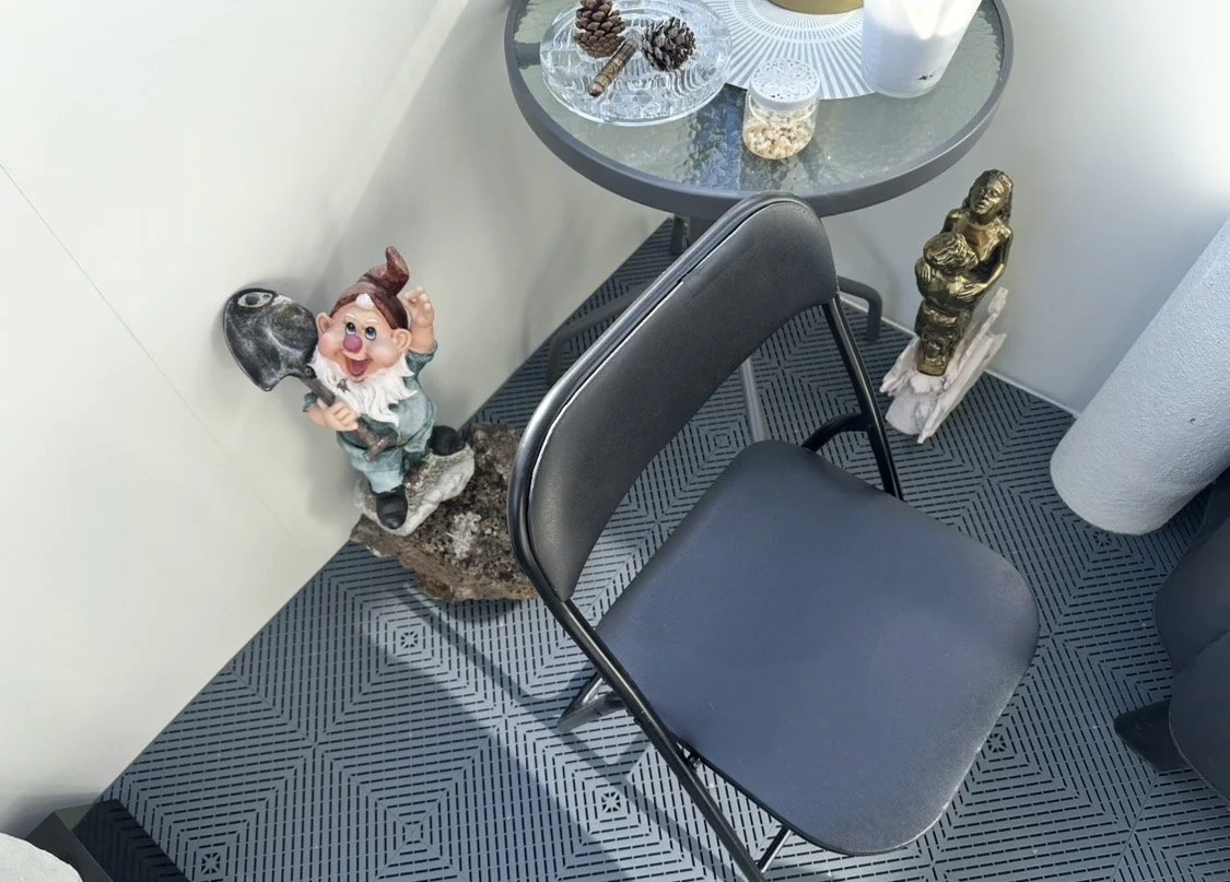 balcony with a decorative gnome figurine next to a black chair in a corner near a glass table, all on swisstrax tile floor.