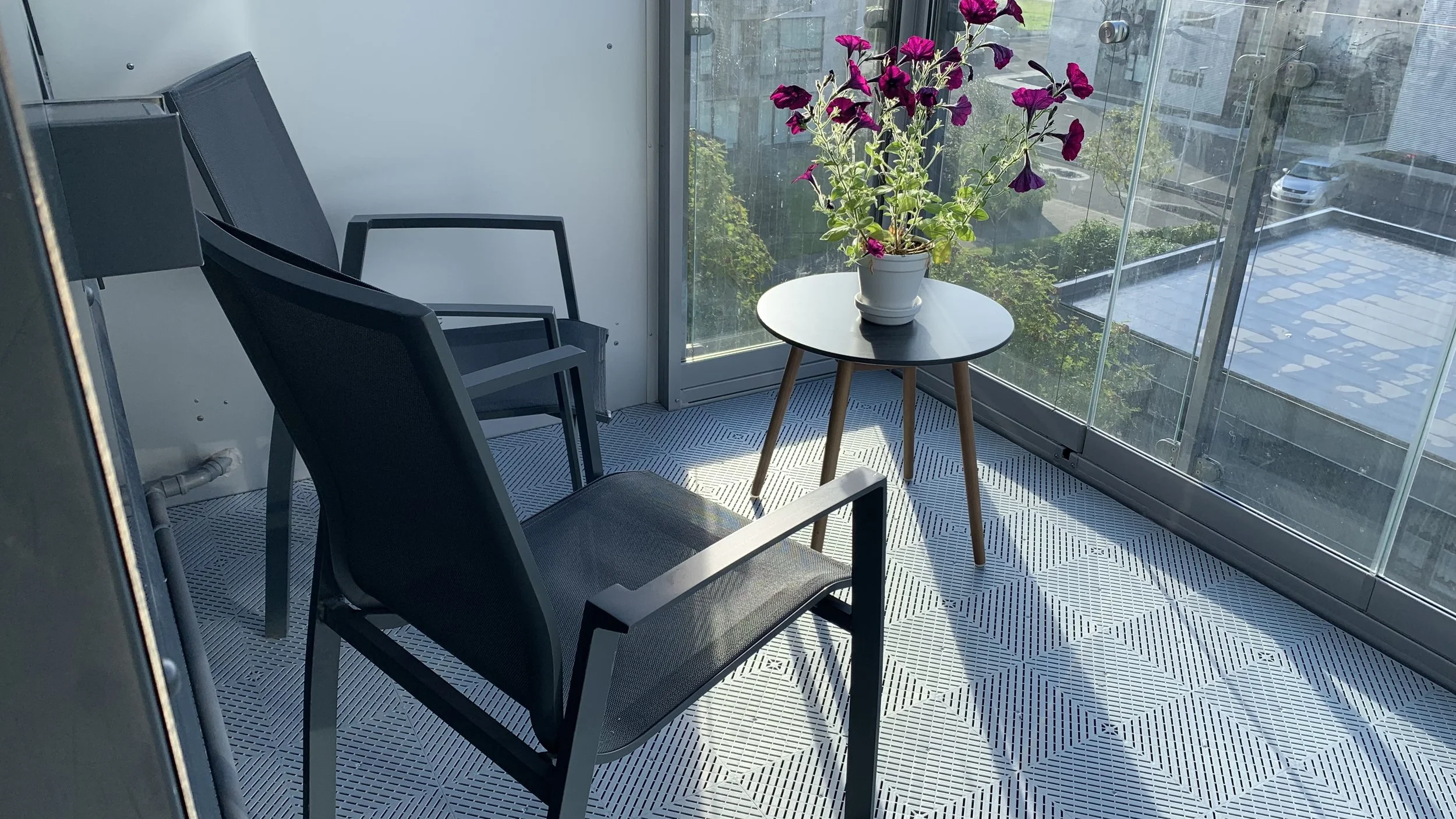 A small balcony with two black chairs, a round black table holding a potted purple flower, swisstrax tile floor and a glass railing with an urban view.