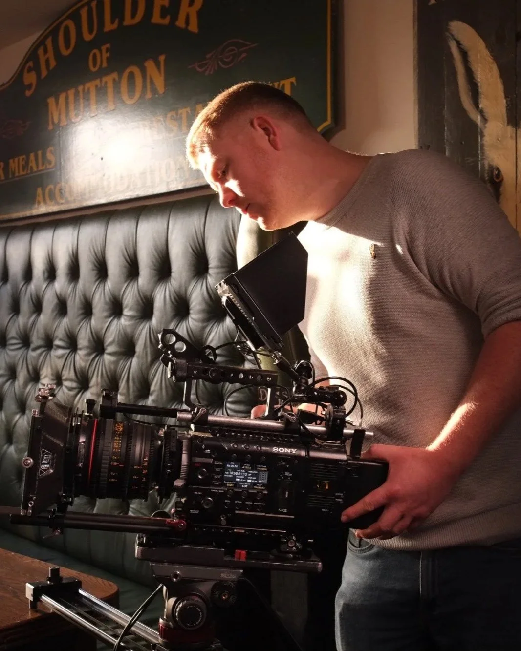 A man operating a professional video camera on a set with a poster in the background that reads 'Shoilder of Mutton'.