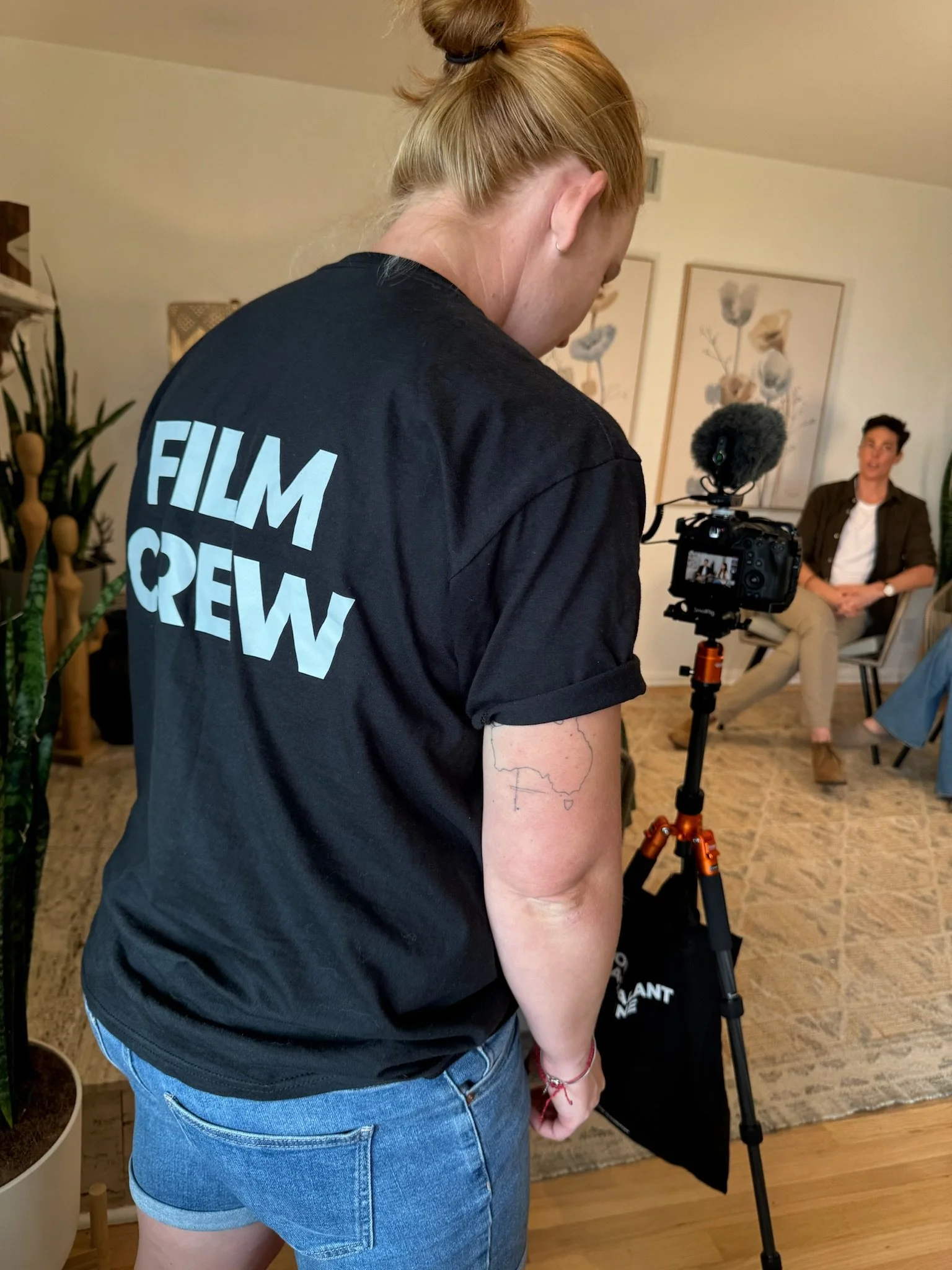 A person with red hair in a bun and a black T-shirt that says 'FILM CREW' on the back is standing in front of a camera on a tripod. The person has a tattoo on their left arm and is wearing denim shorts.