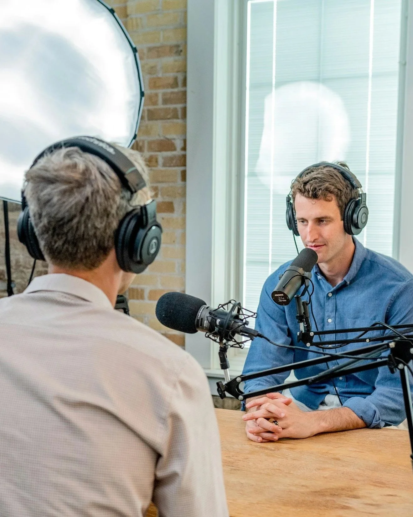 Two men are recording a podcast or radio show in a studio. They are wearing headphones and sitting at a table with microphones. One man with glasses is facing away from the camera, and the other man with dark hair is facing towards the camera, speaking into a microphone.