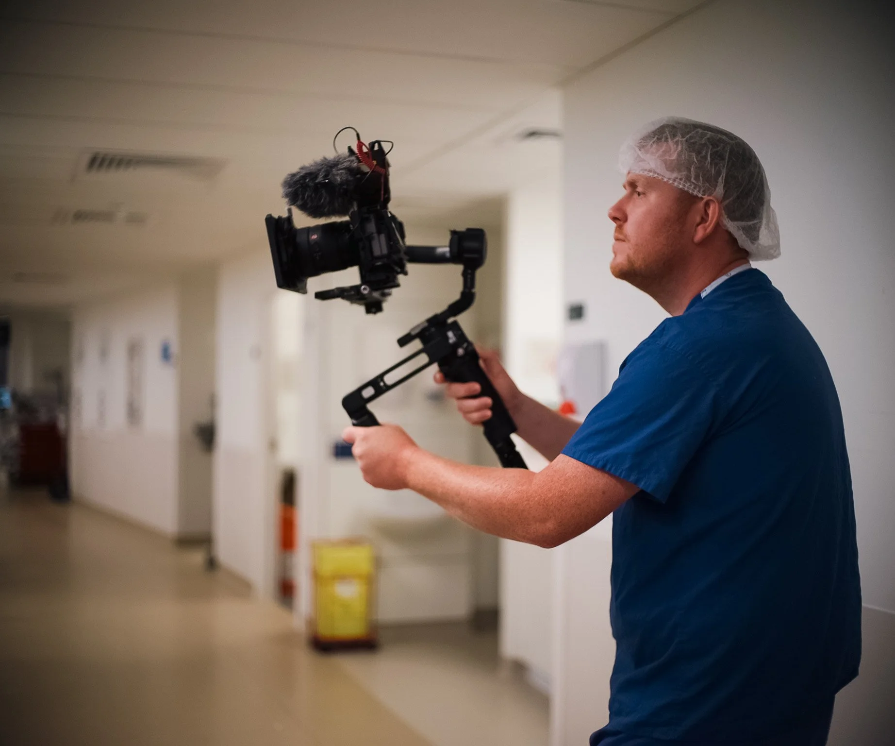 A man wearing a hairnet and scrubs operating a professional camera in an indoor hallway, possibly in a hospital or medical facility.