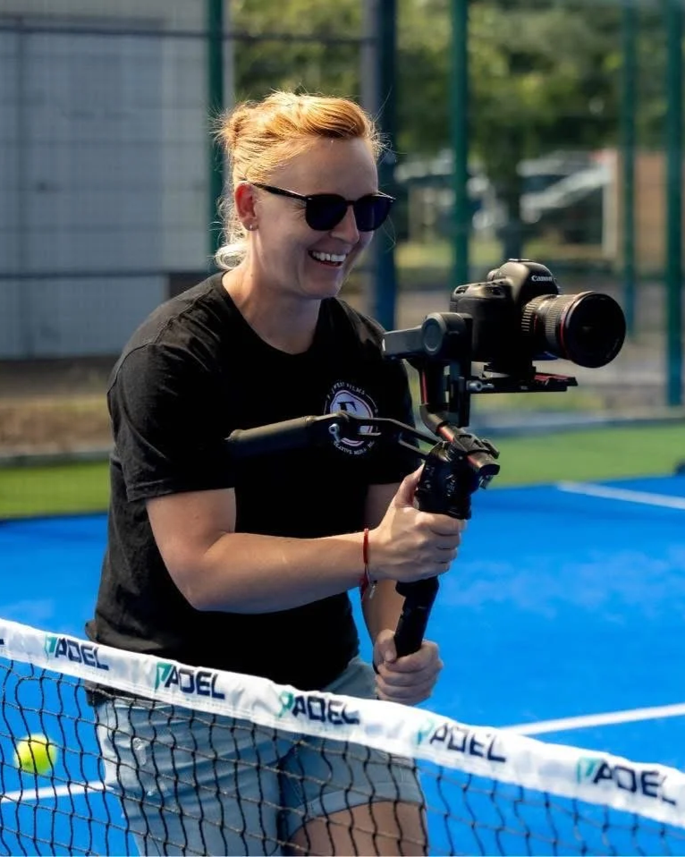 A woman with blonde hair, wearing sunglasses, a black t-shirt, and gray shorts, is holding a camera stabilizer with a camera attached. She is standing next to a tennis net on a blue tennis court, smiling. There is a green fence and trees in the background.