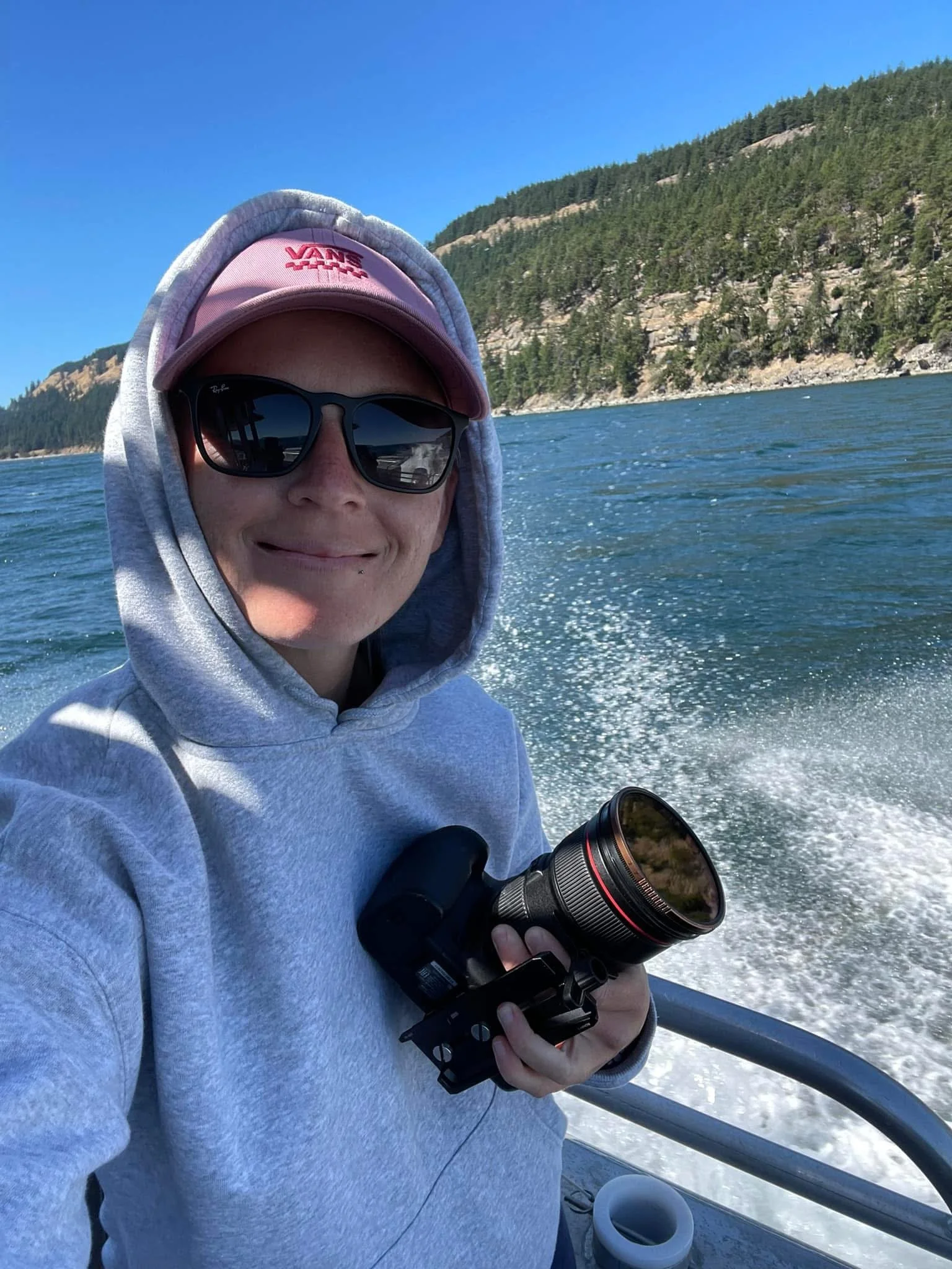 Person wearing sunglasses, a pink cap, and a hoodie, holding a camera with a scenic water and mountain background.