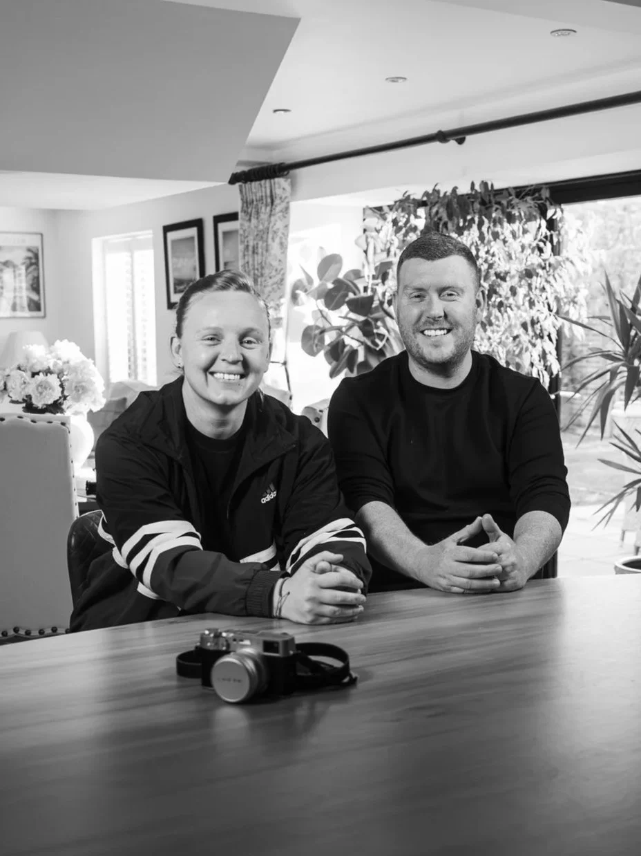 Two smiling men sitting at a wooden table, with a camera on the table, in a well-lit room with large windows and plants in the background.