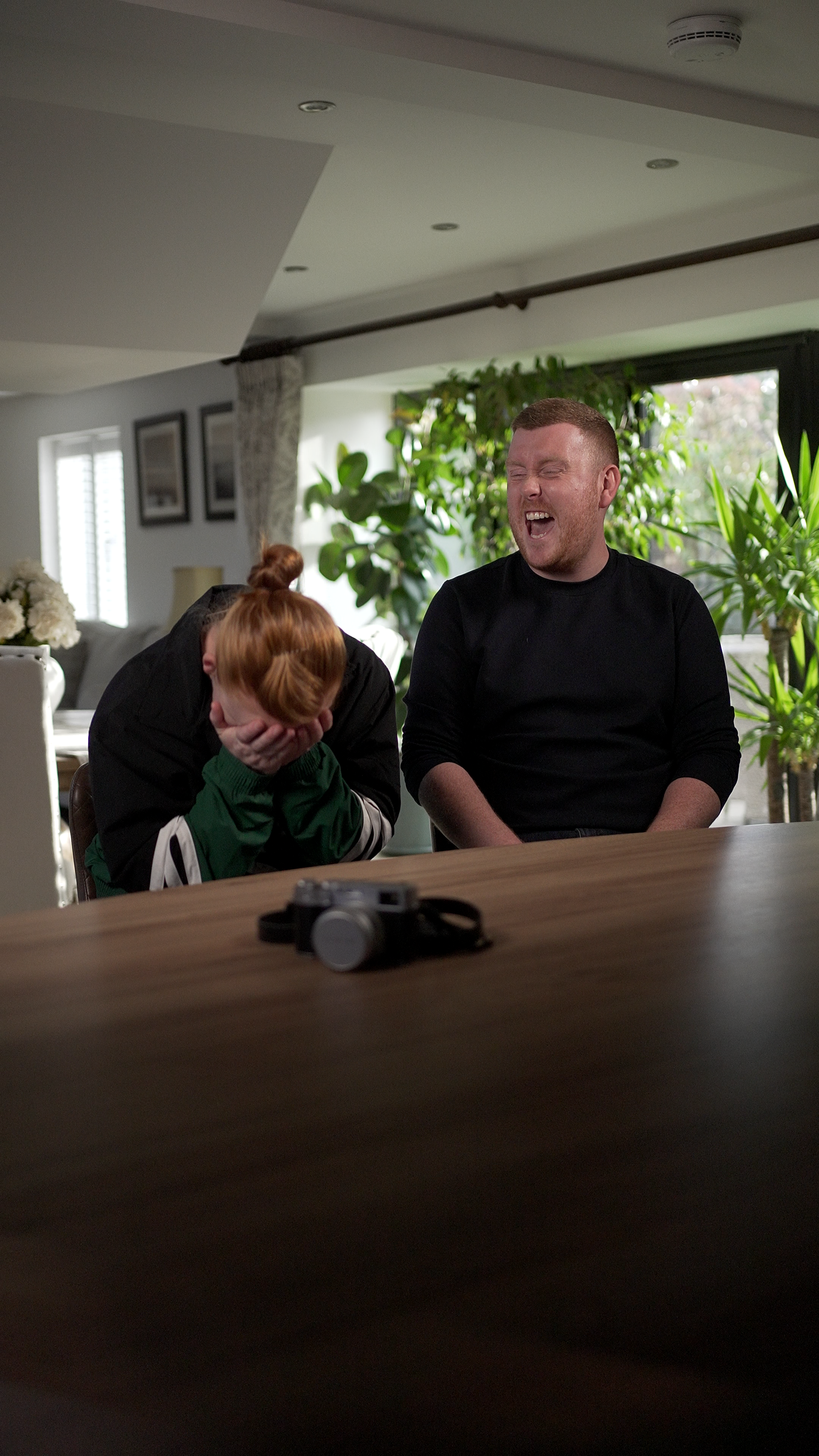 A man and a woman are seated at a table in a bright, plant-filled room. The woman has red hair and is covering her face with her hands, while the man is smiling or laughing with his eyes closed.
