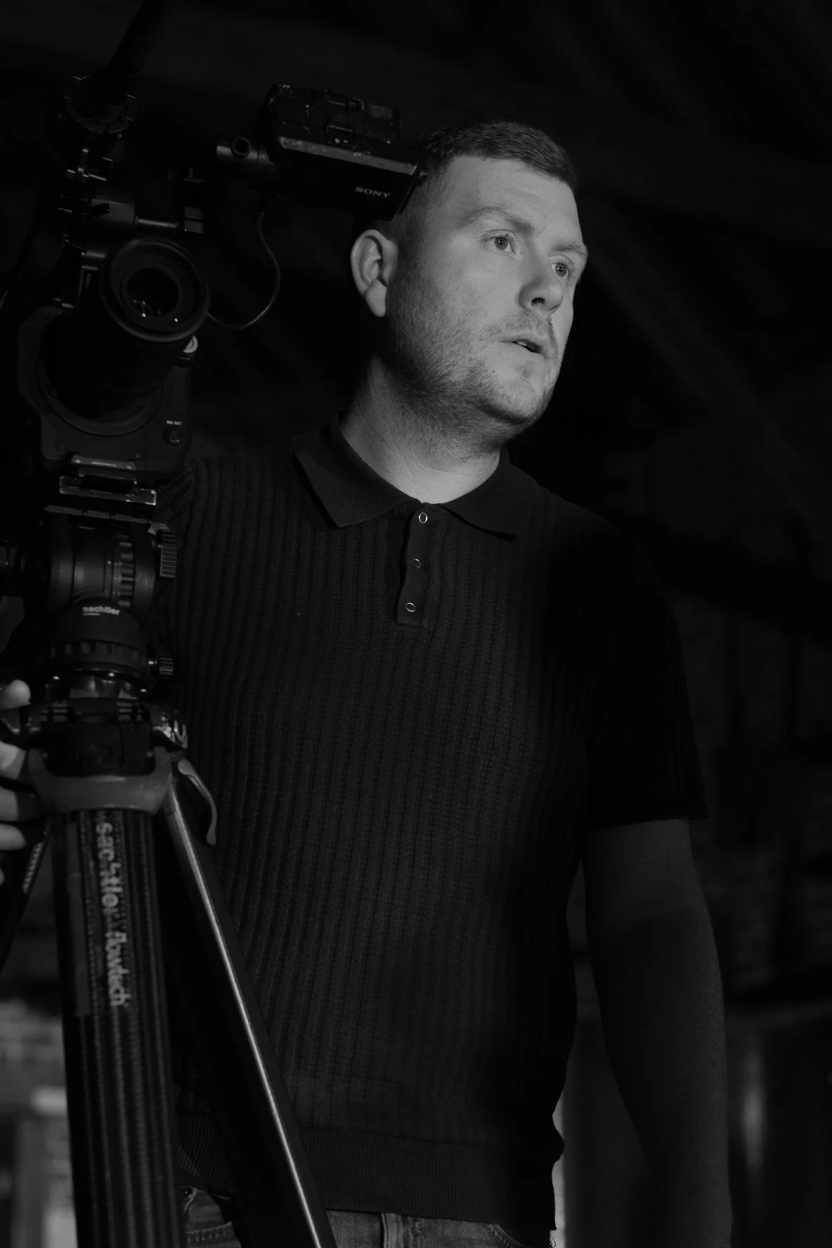A man with short hair wearing a dark, short-sleeved polo shirt, standing next to a professional video camera on a tripod.