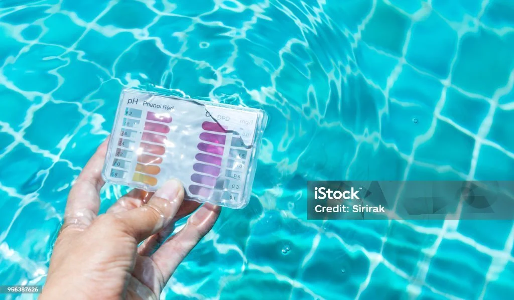 Person holding a pool water test kit checking pH and chlorine levels against a color chart, with a swimming pool in the background.