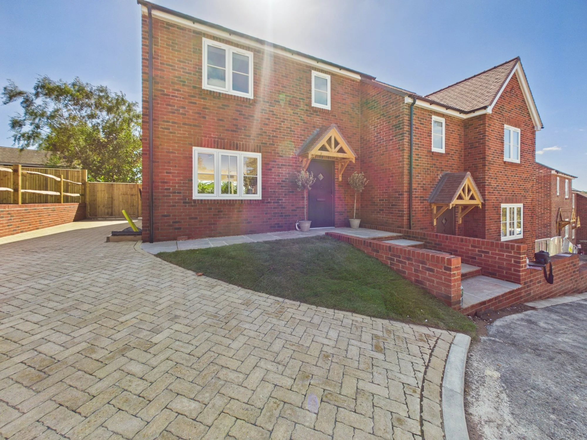 A residential brick house with a paved driveway, two small trees in pots near the front door, and a clear blue sky with sunlight reflecting glare.