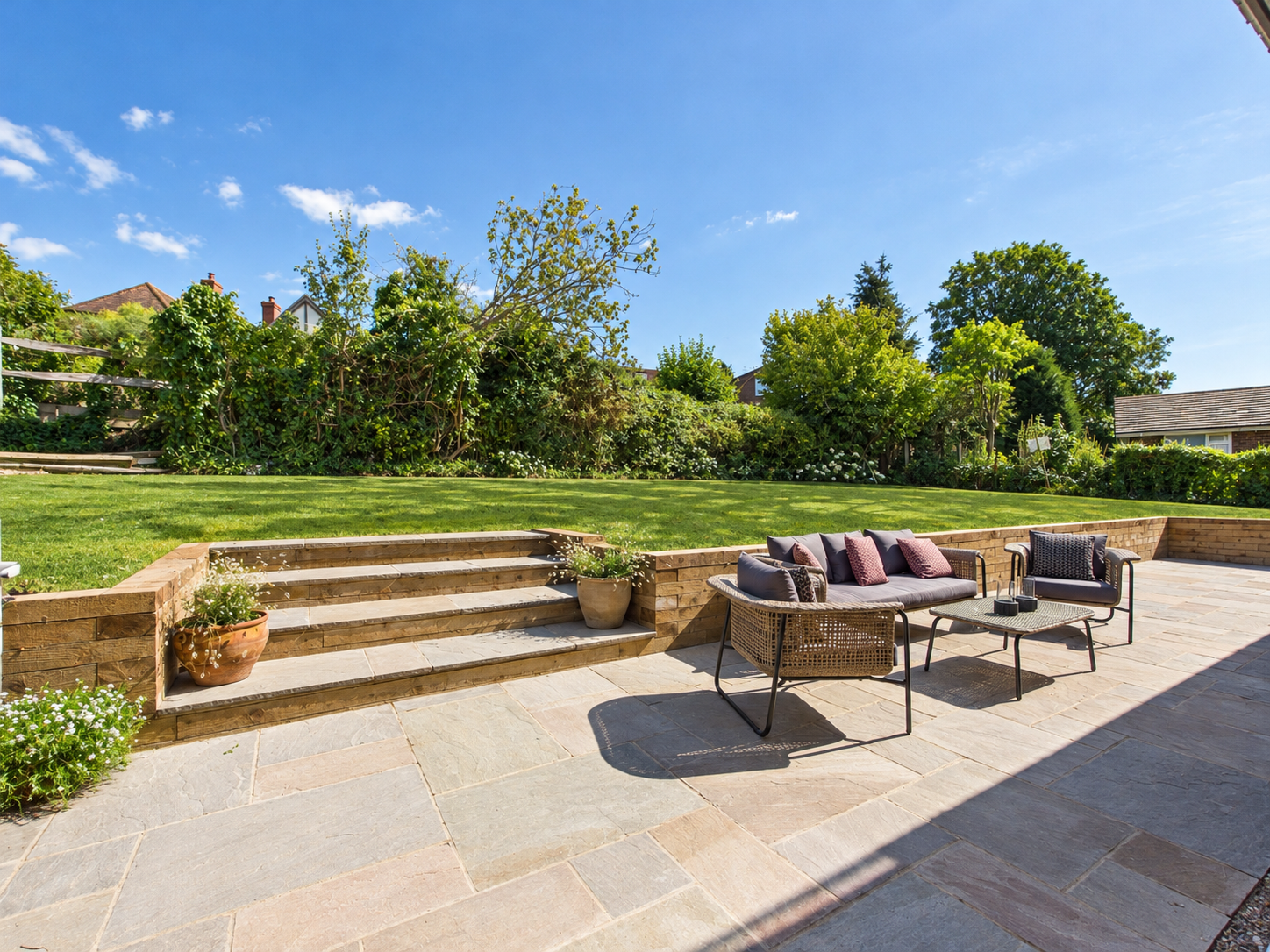A backyard patio with outdoor seating, potted plants, and a grassy yard with trees under a blue sky.