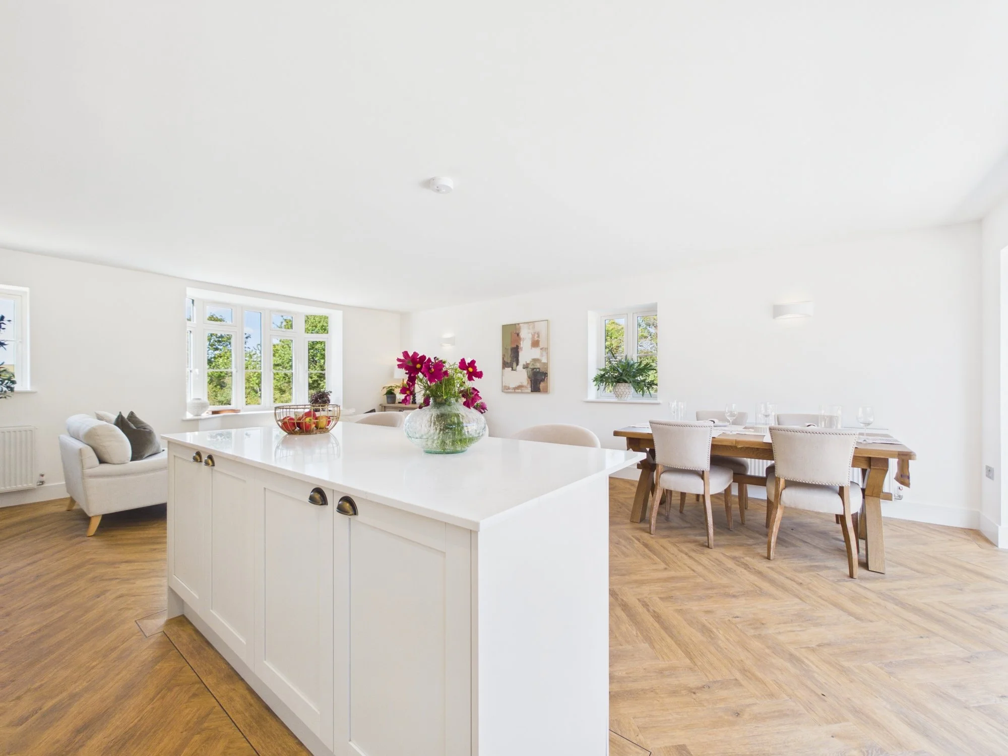 Bright and spacious living and dining area with white walls, large windows, hardwood floors, a beige sofa, dining table with chairs, a decorative painting, and potted plants.