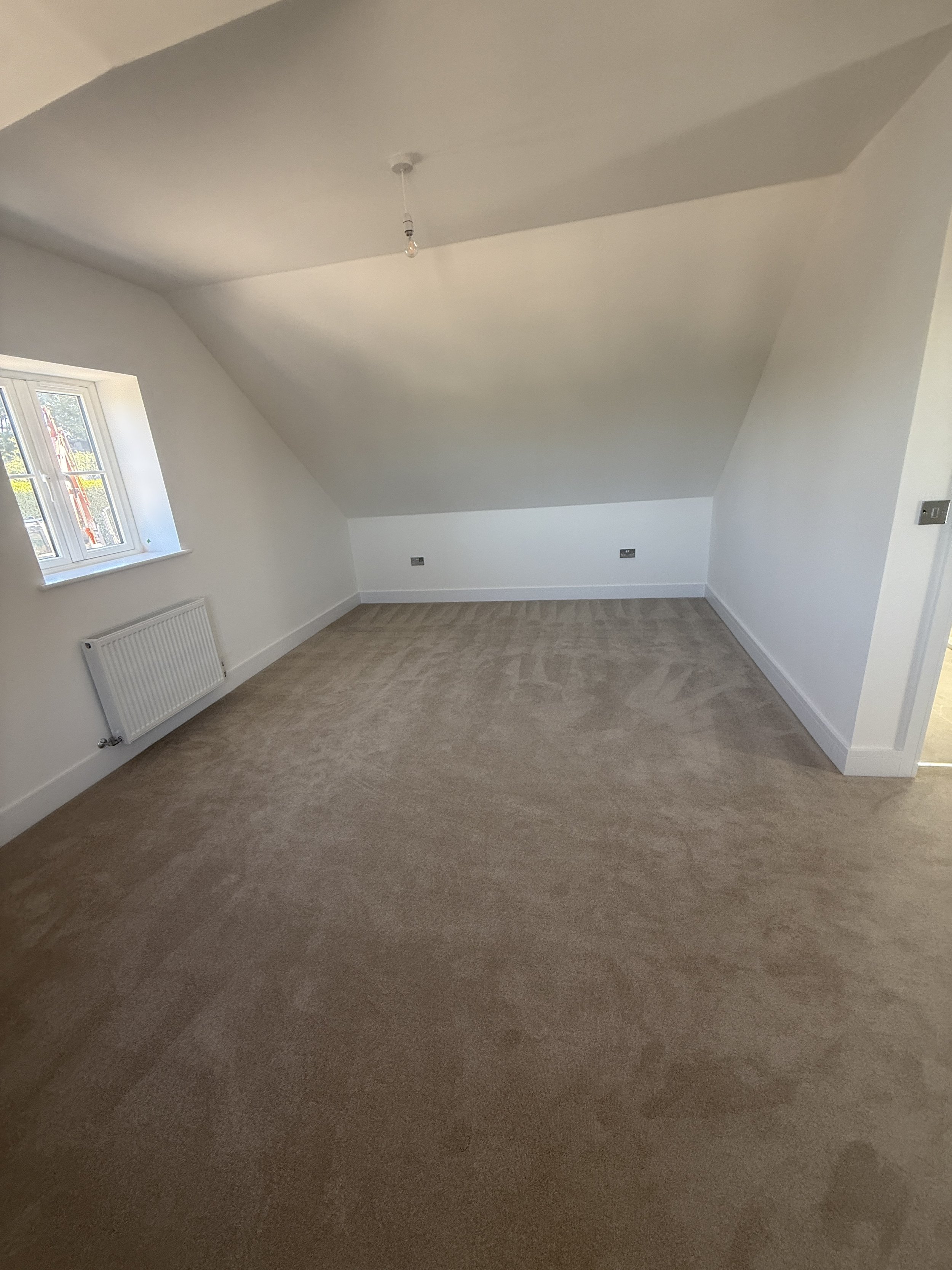 An empty room with light brown carpet, white walls, a small window, ceiling light fixture, and electrical outlets.