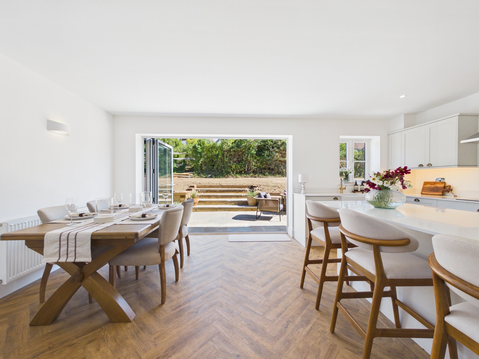 Bright, modern dining area with white walls, wooden flooring, a wooden dining table with seating, and large open sliding doors leading to an outdoor space with greenery.