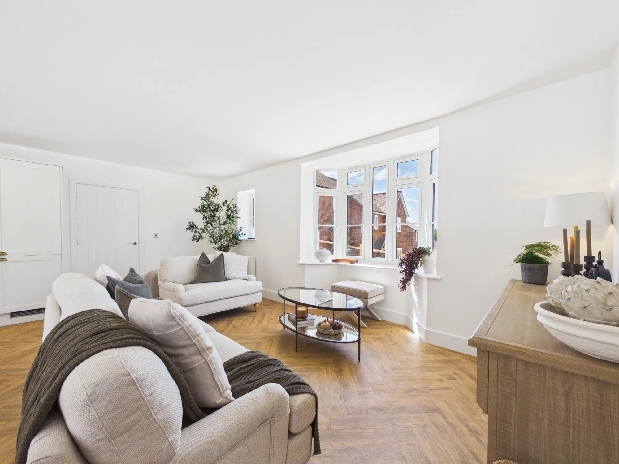 Bright living room with white walls, large bay window, cozy beige sofas with gray pillows, wooden flooring, and various indoor plants.