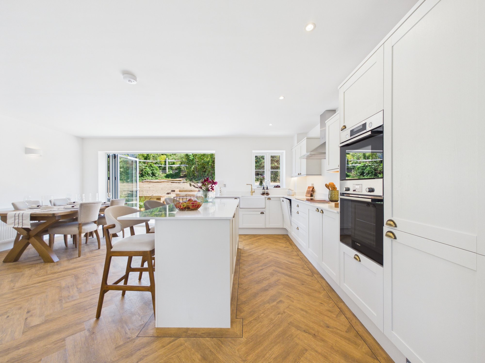 A bright white kitchen with a central island, white cabinets, and modern appliances, adjacent to a dining area with a wooden table and beige chairs, overlooking an outdoor yard through open glass doors.