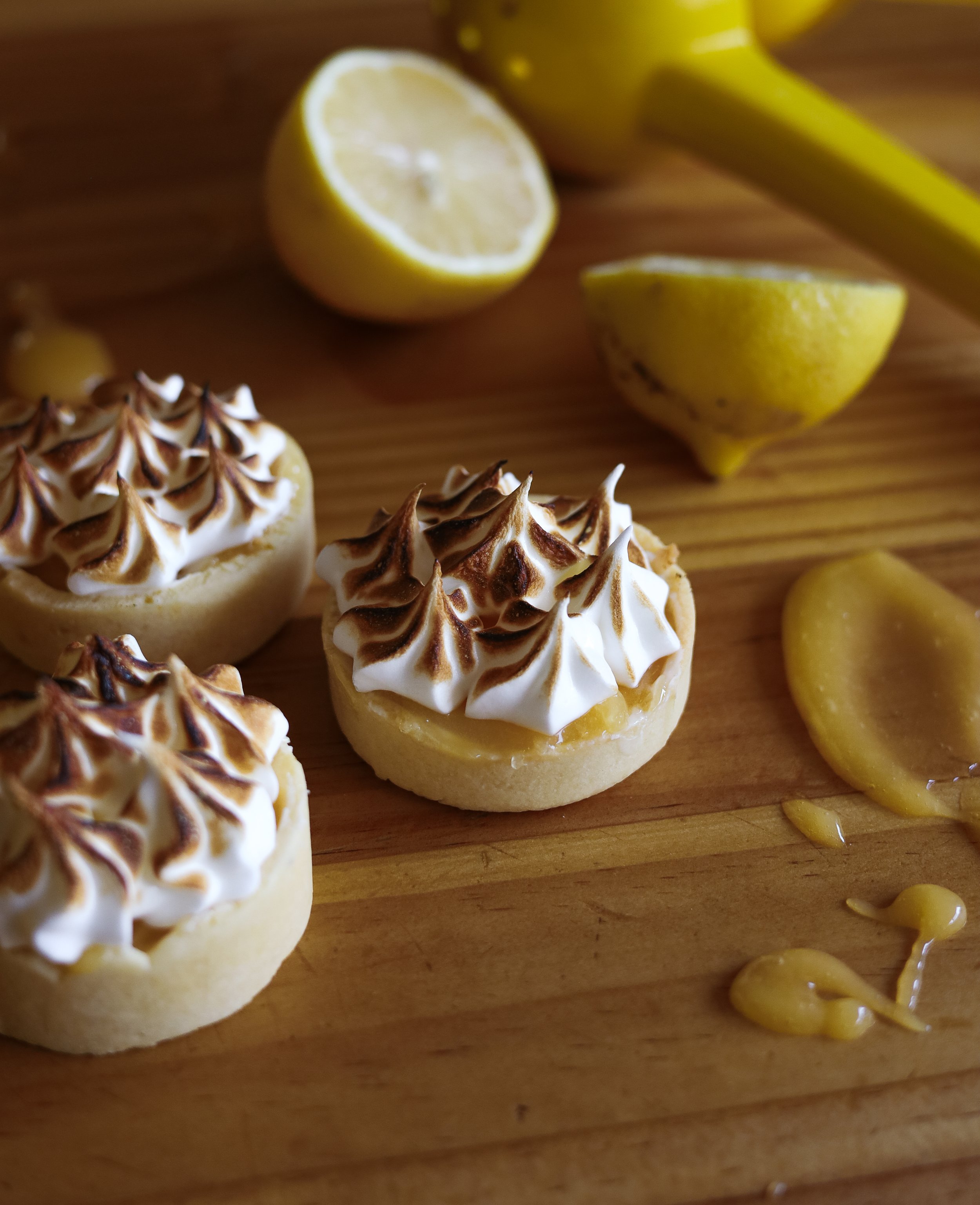 Lemon tarts with toasted meringue on a wooden surface with lemon slices and a yellow lemon juicer in the background.