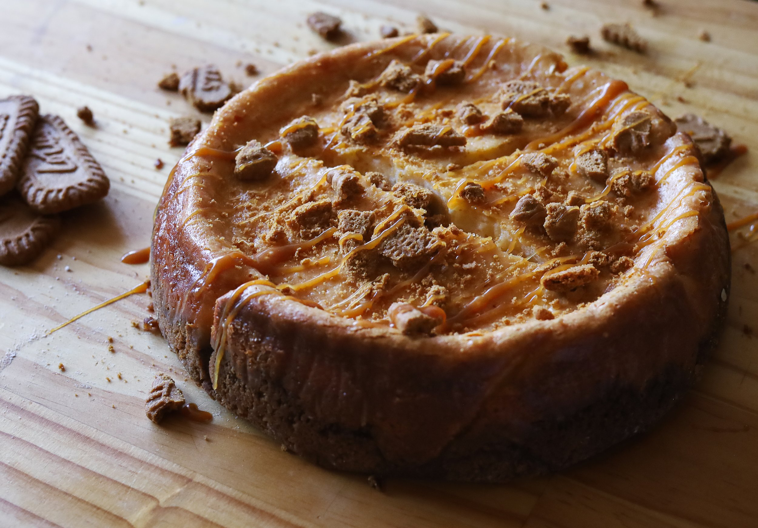 A Baked Biscoff and Caramel Cheesecake with caramel drizzle and cookie pieces on top, placed on a wooden surface alongside a few Biscoff cookies.