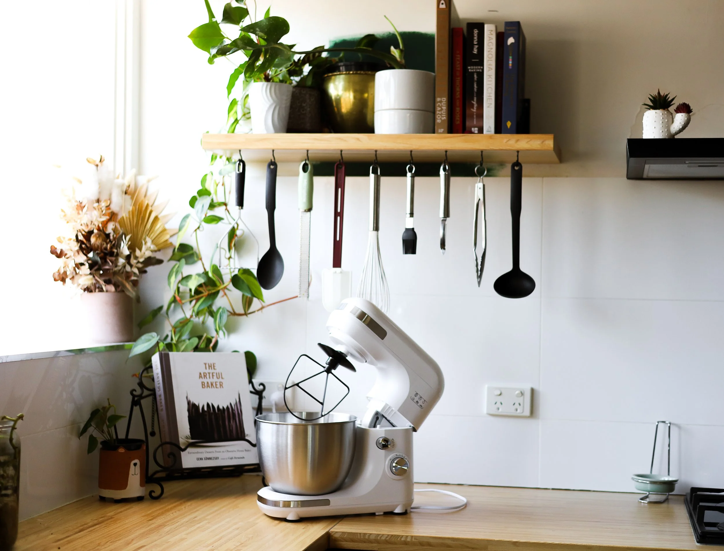 Kitchen countertop with a white stand mixer, open cookbook titled 'The Artful Baker', potted plants, and hanging kitchen utensils on a wooden shelf. White walls with an electrical outlet, and a window on the left side.