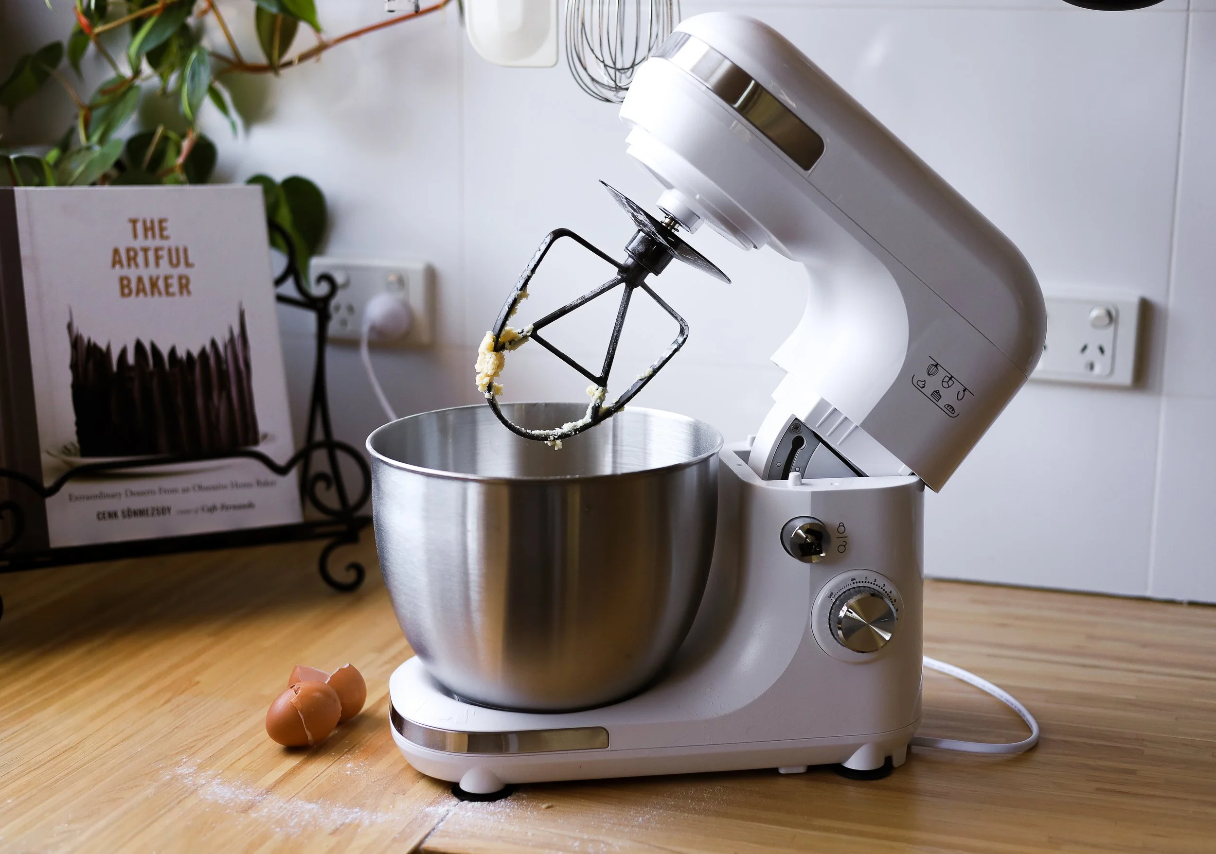 Kitchen countertop with a stand mixer, metal mixing bowl, and eggs, with a cookbook titled "The Artful Baker" on a stand.