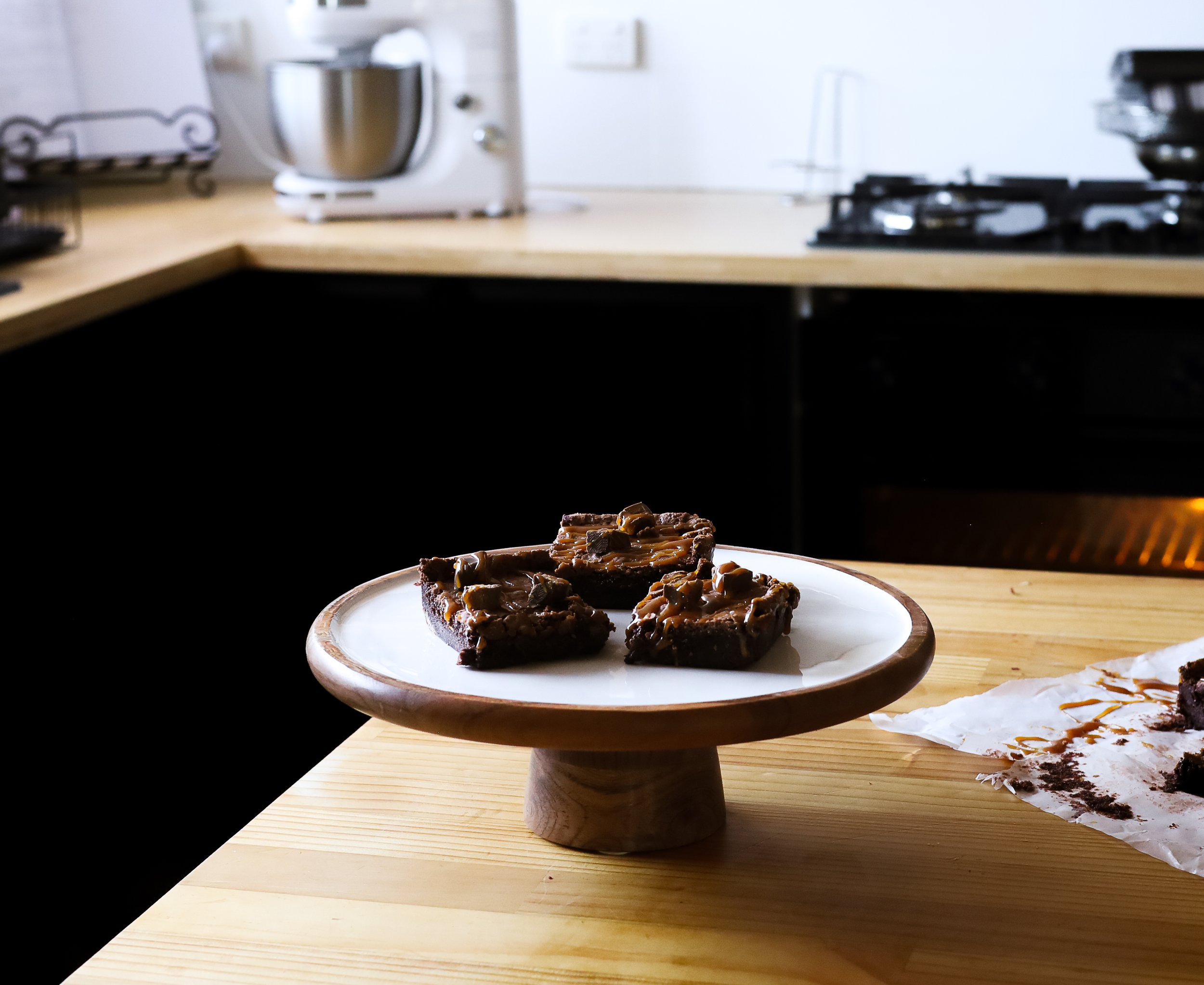 Three pieces of chocolate brownie topped with Caramel on a white plate, placed on a wooden cake stand on a wooden table in a kitchen.