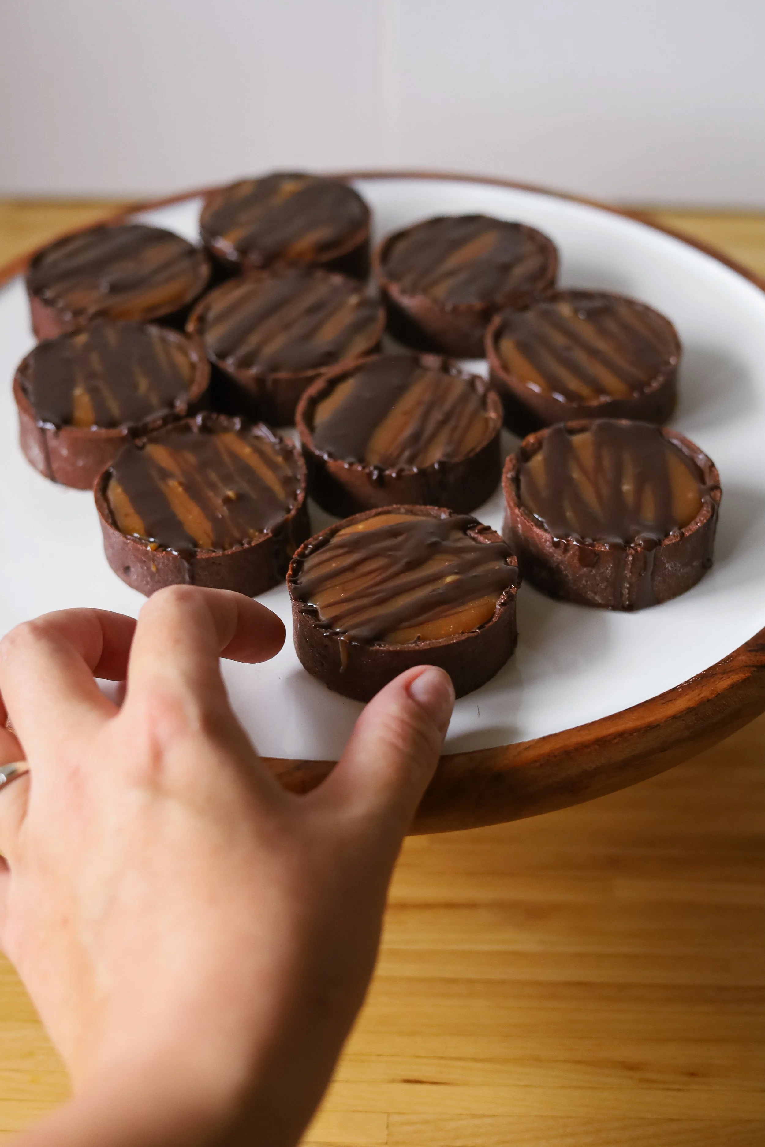 Hand reaching for a chocolate tart with caramel and chocolate drizzles on a white plate.