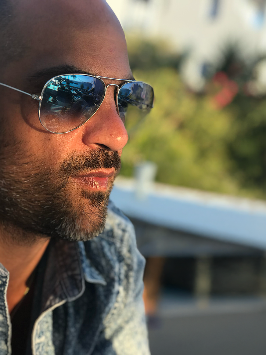 Close-up side profile of a man wearing reflective sunglasses and a denim jacket outdoors during the day.