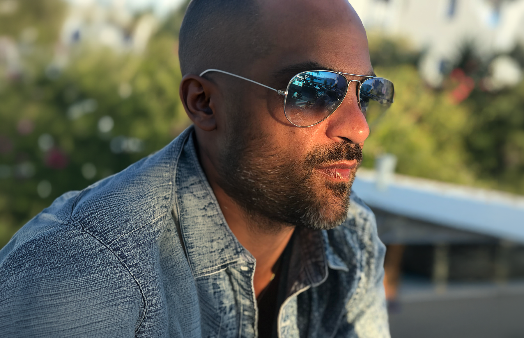 Close-up of a man with short hair, sunglasses, and a denim jacket, outdoors with greenery in the background.