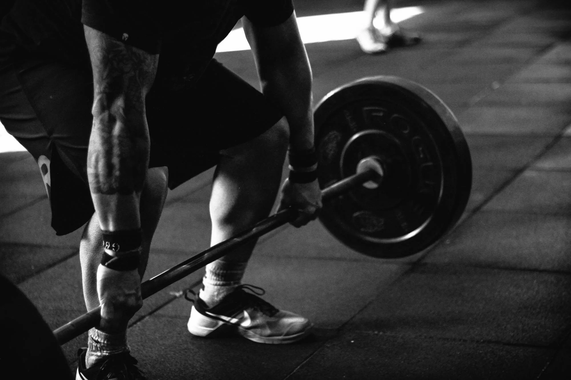 A person is in a gym, lifting a barbell with a weight plate on it. The scene is in black and white, with the person's arms and legs visible, wearing shorts, sneakers, and socks. The background is slightly out of focus.