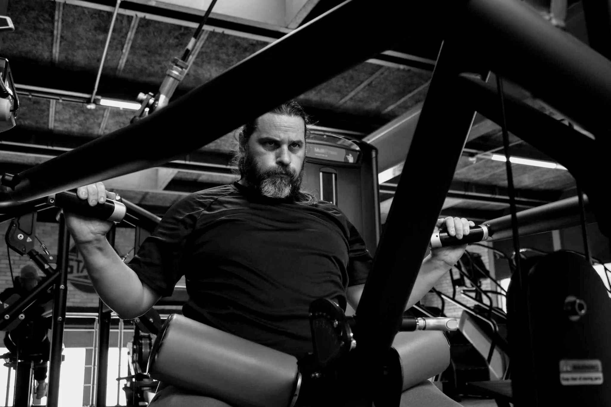 A man with a beard sitting on a rowing machine in a gym, holding onto the handle with both hands and looking forward, surrounded by gym equipment and open ceiling with visible pipes.