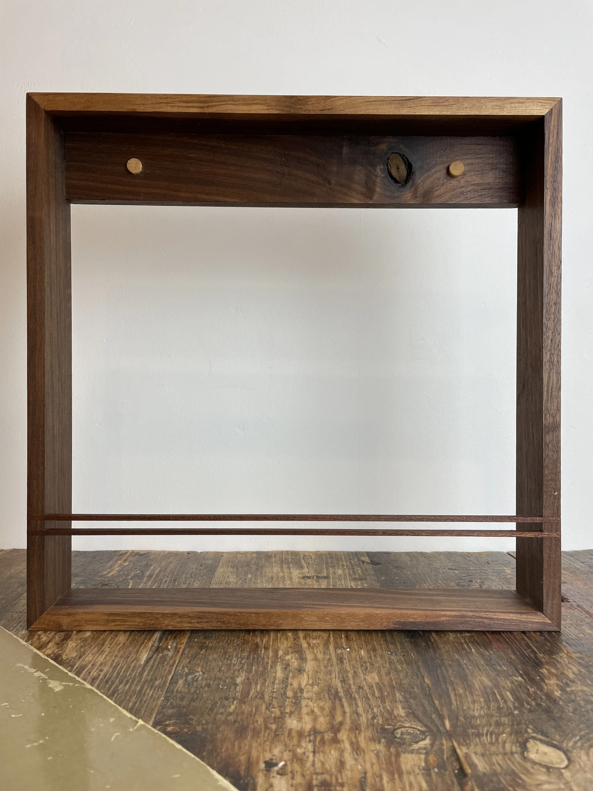 A wooden frame shelf with two small wooden pegs inside, set against a plain white wall.
