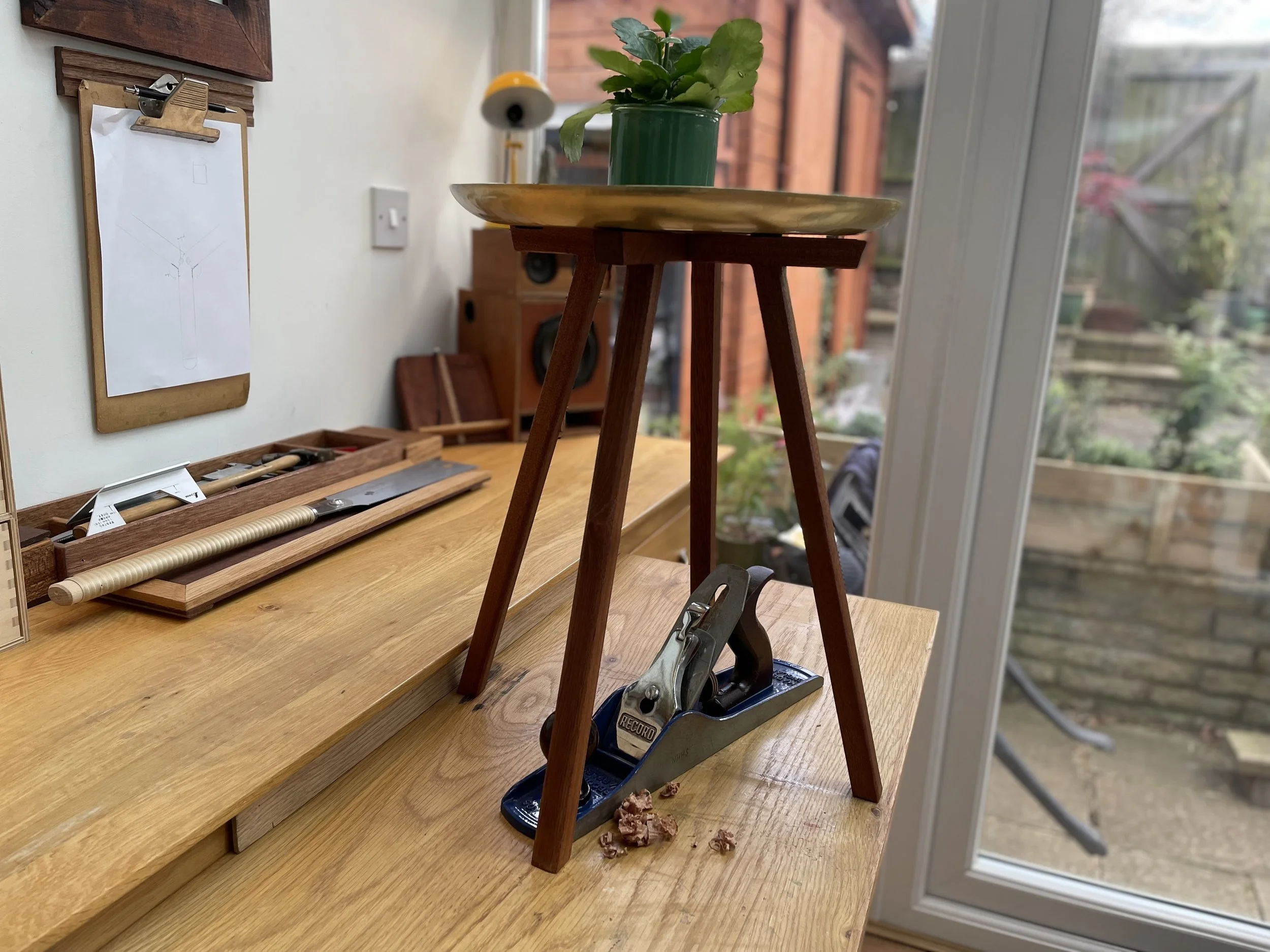 Wooden stool with a potted plant in a green pot on top, set on a light wooden table inside a room with a large glass door. Various woodworking tools and scraps are scattered on the table. In the background, a white wall with a clipboard holding sketc
