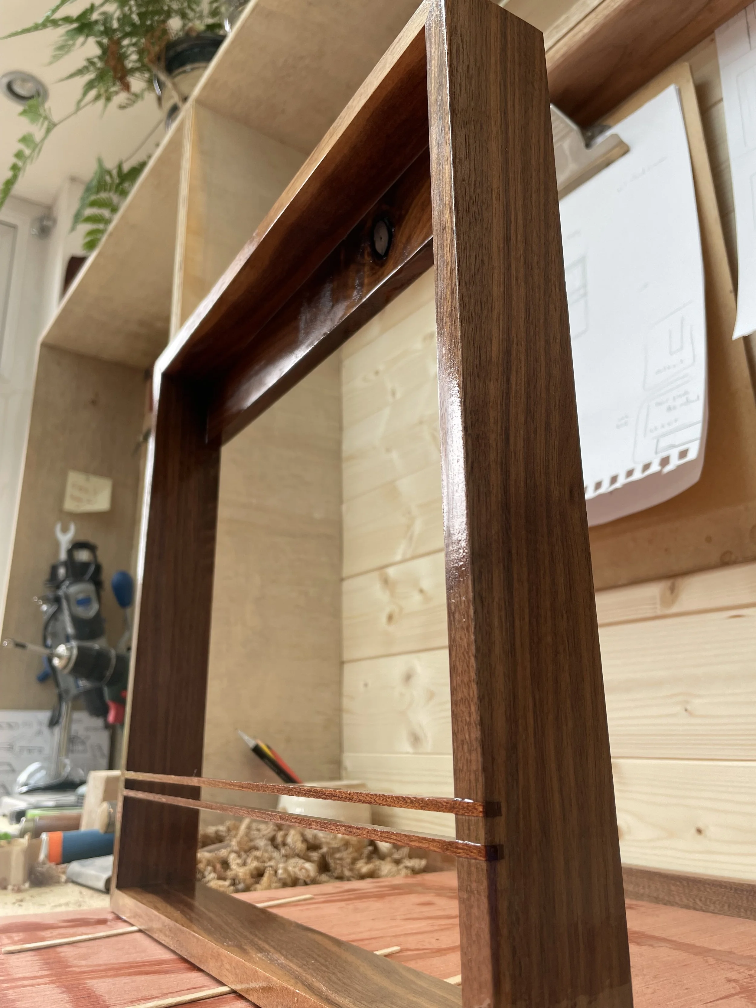 A wooden piece of furniture frame, possibly a bookshelf or shelf, being assembled on a wooden table in a workshop. A screwdriver and some tools are visible in the background.