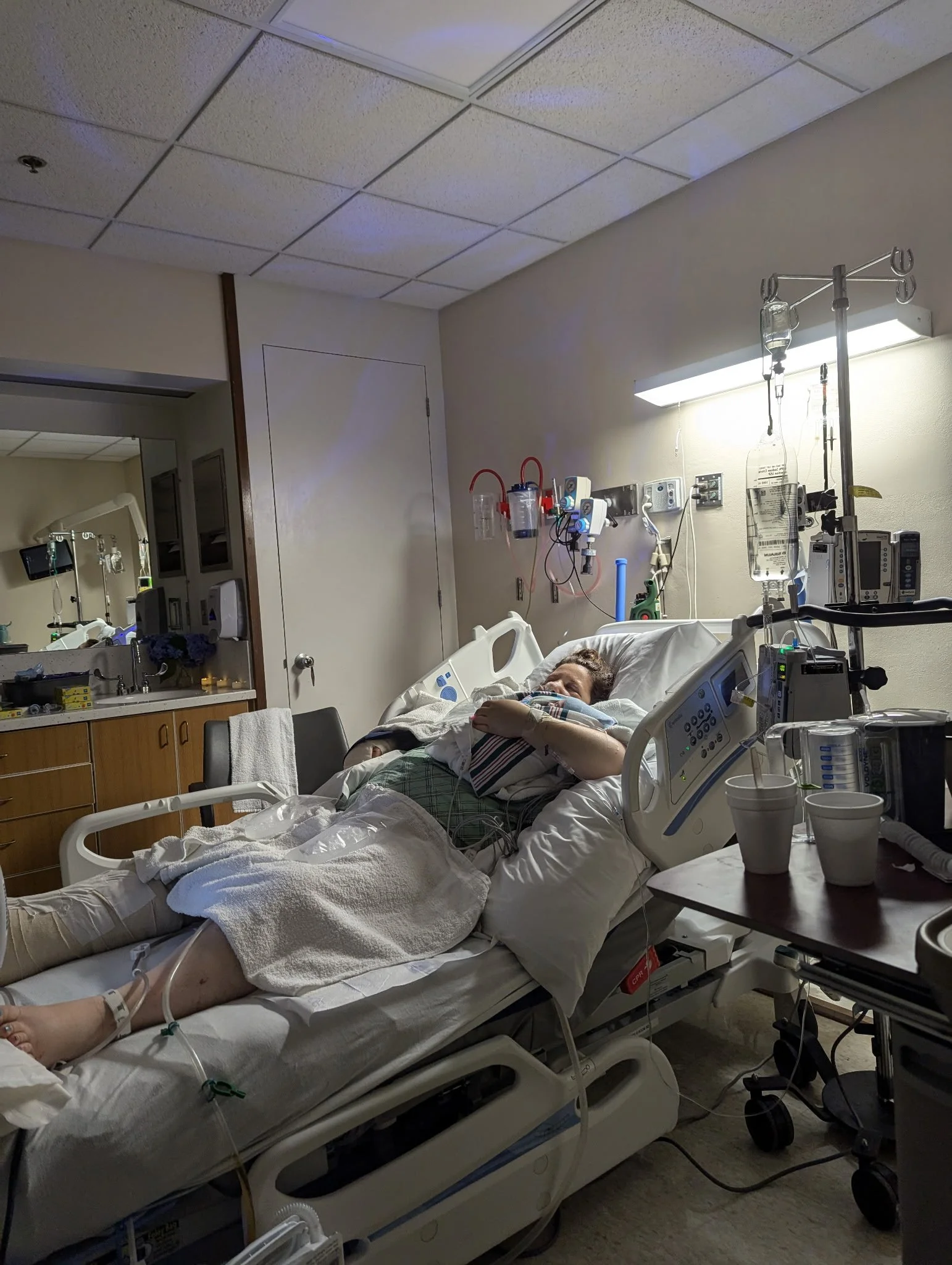 A person lying in a hospital bed connected to medical equipment, with IV bags hanging, in a hospital room.