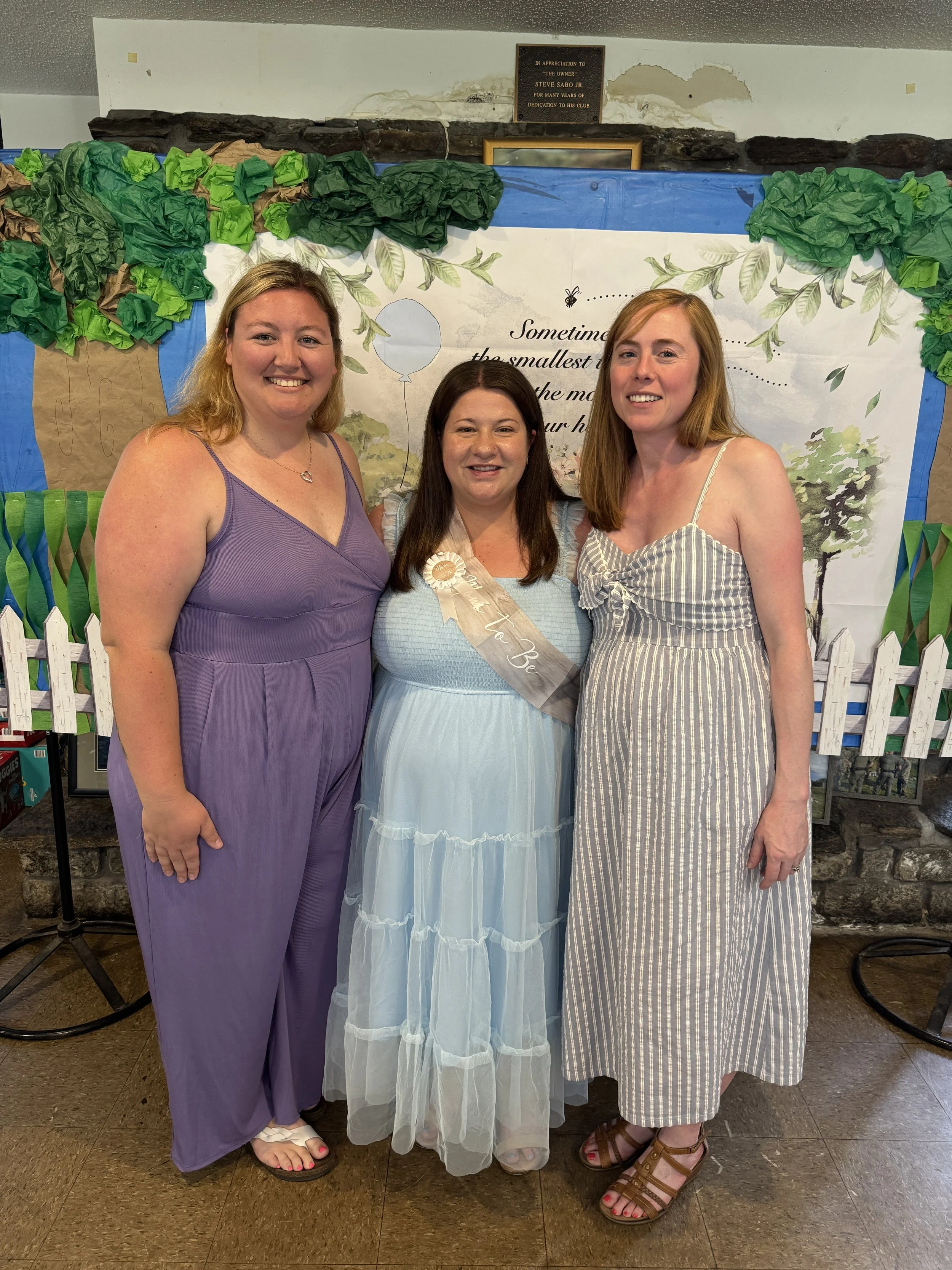 Three women standing together at a celebration, with a decorated backdrop featuring trees and text, one woman wearing a sash and a dress, the others in summer outfits.
