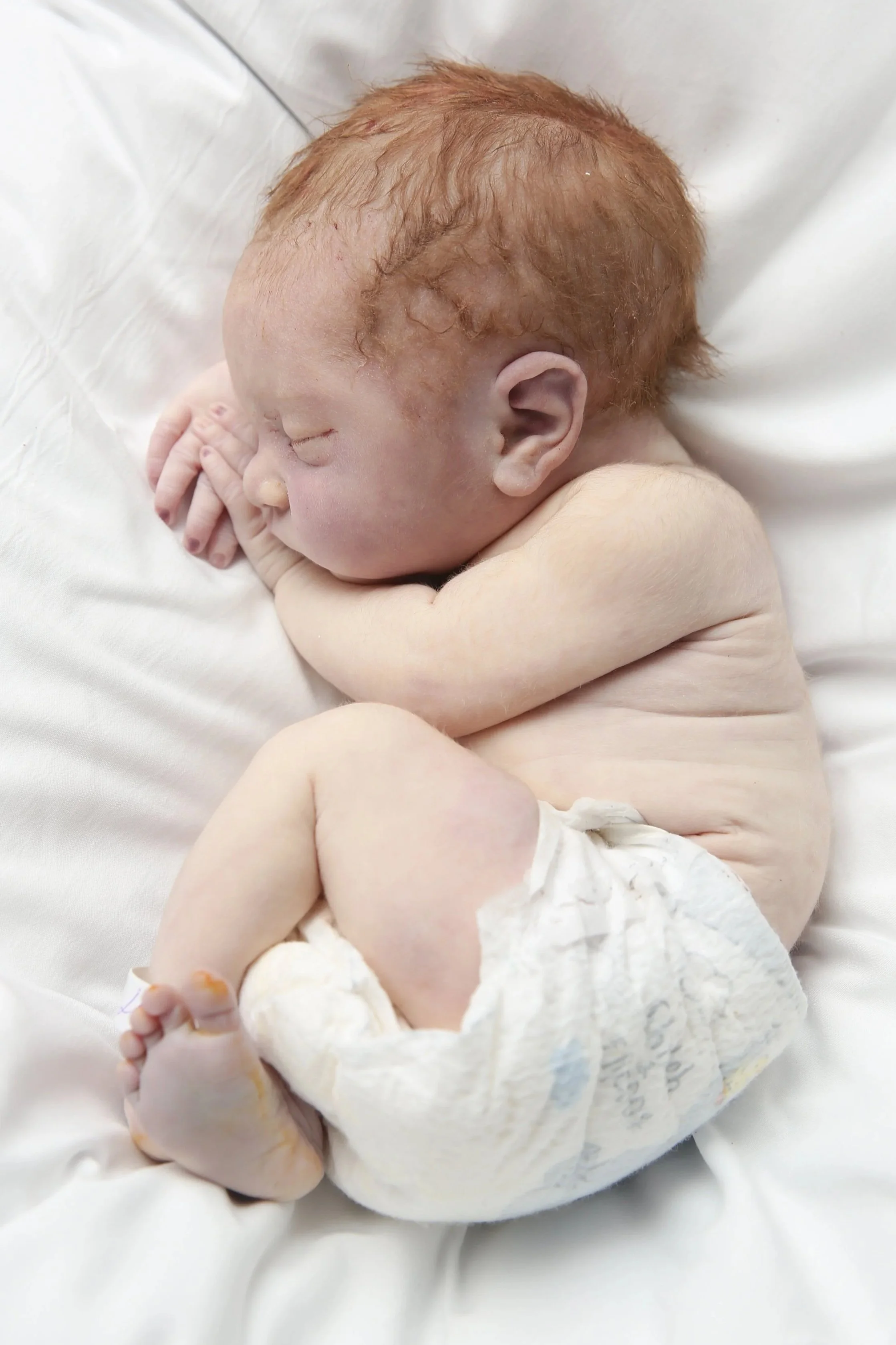 A sleeping newborn baby with red hair, lying on a white sheet, curled up on their side with arms and legs close to their body, wearing a diaper.