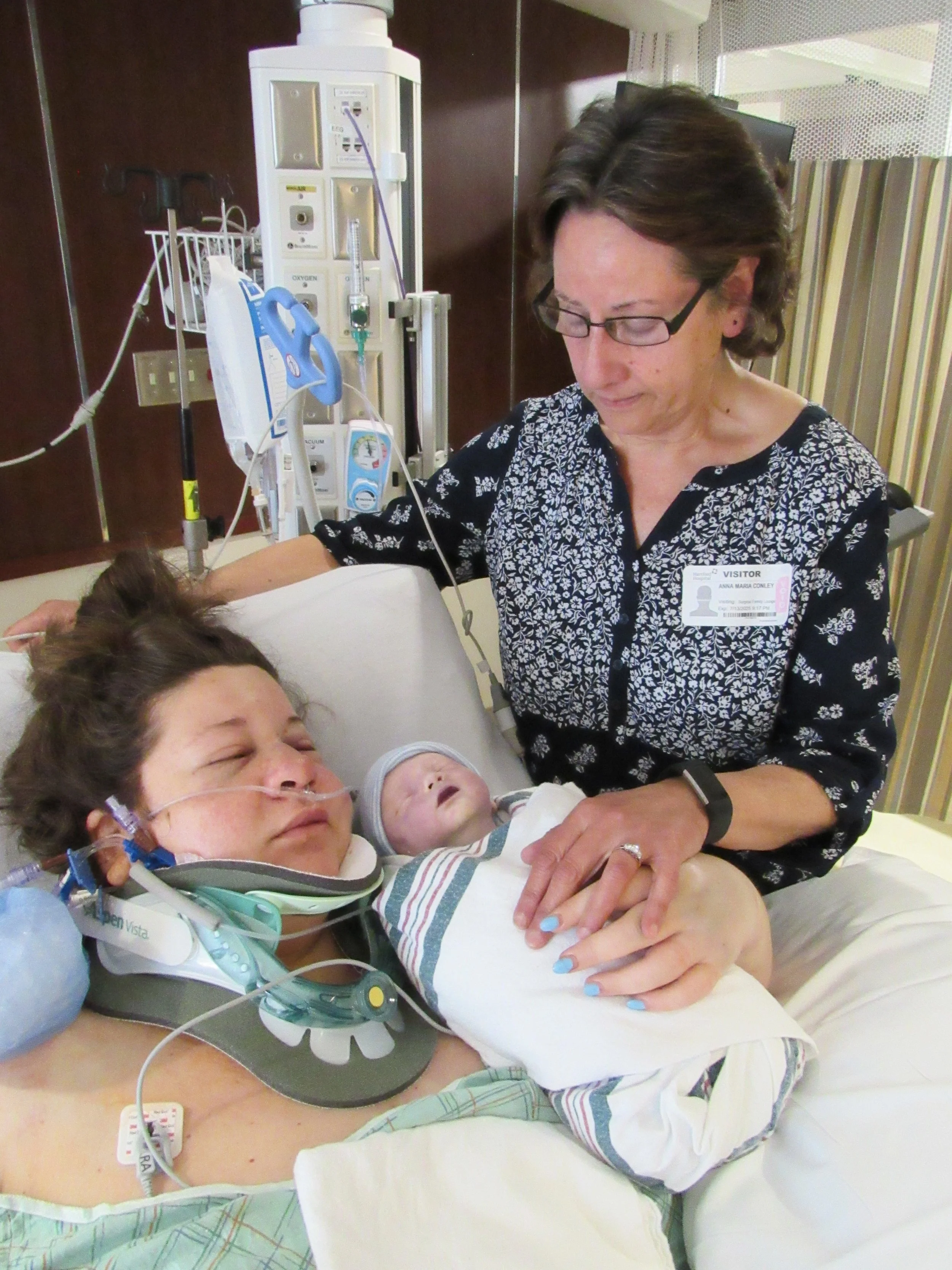 A woman in a hospital bed with medical equipment and an oxygen tube, holding an infant dressed in hospital clothing, while a visitor stands beside her, gently touching her hand.