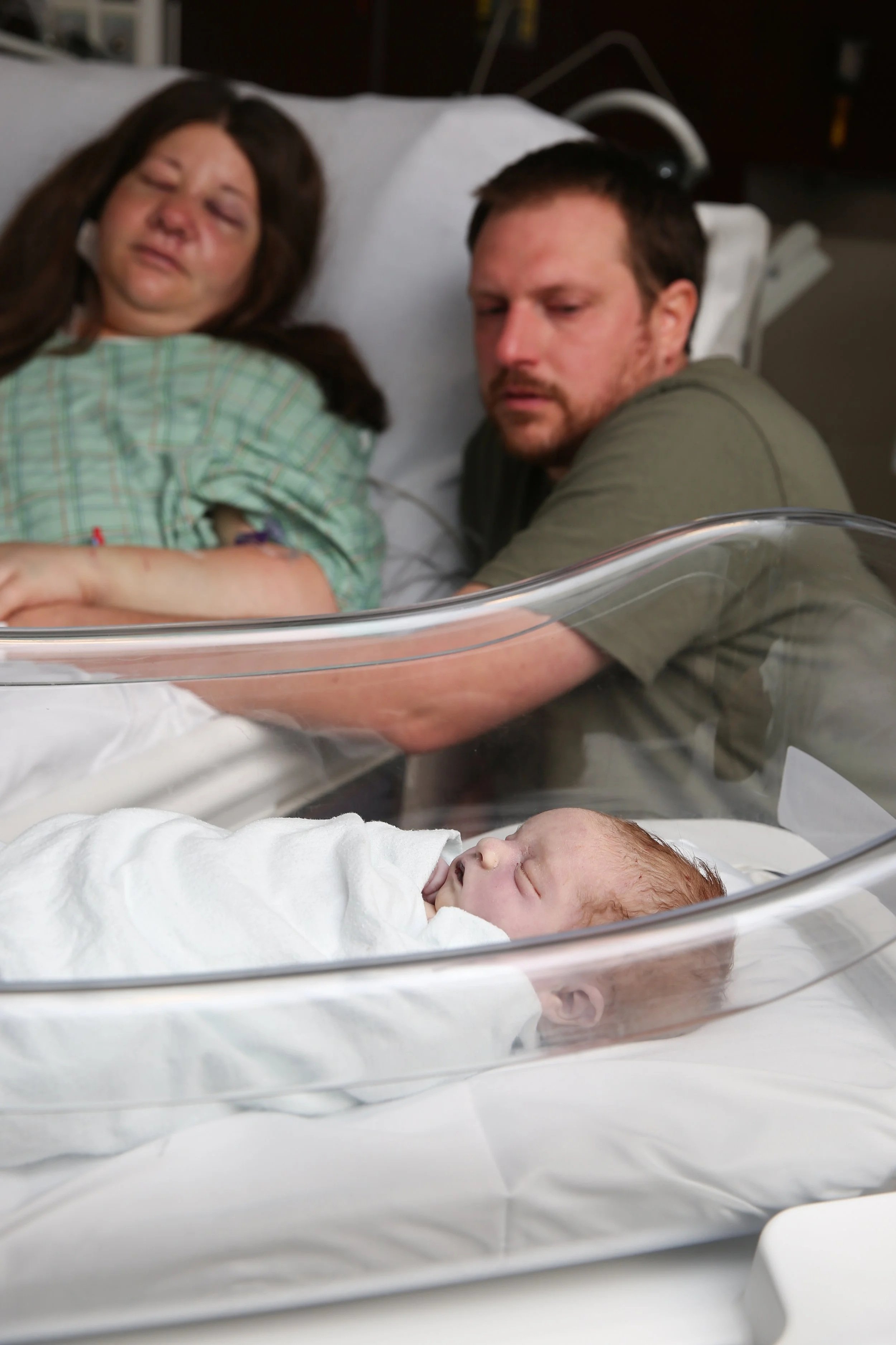A newborn baby sleeping in a clear hospital bassinet, with a mother and father looking on from the hospital bed behind it.
