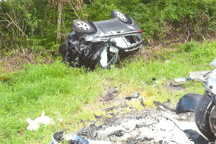 A silver car flipped upside down off the side of the road, surrounded by grass and trees, with significant fire damage and debris scattered around.